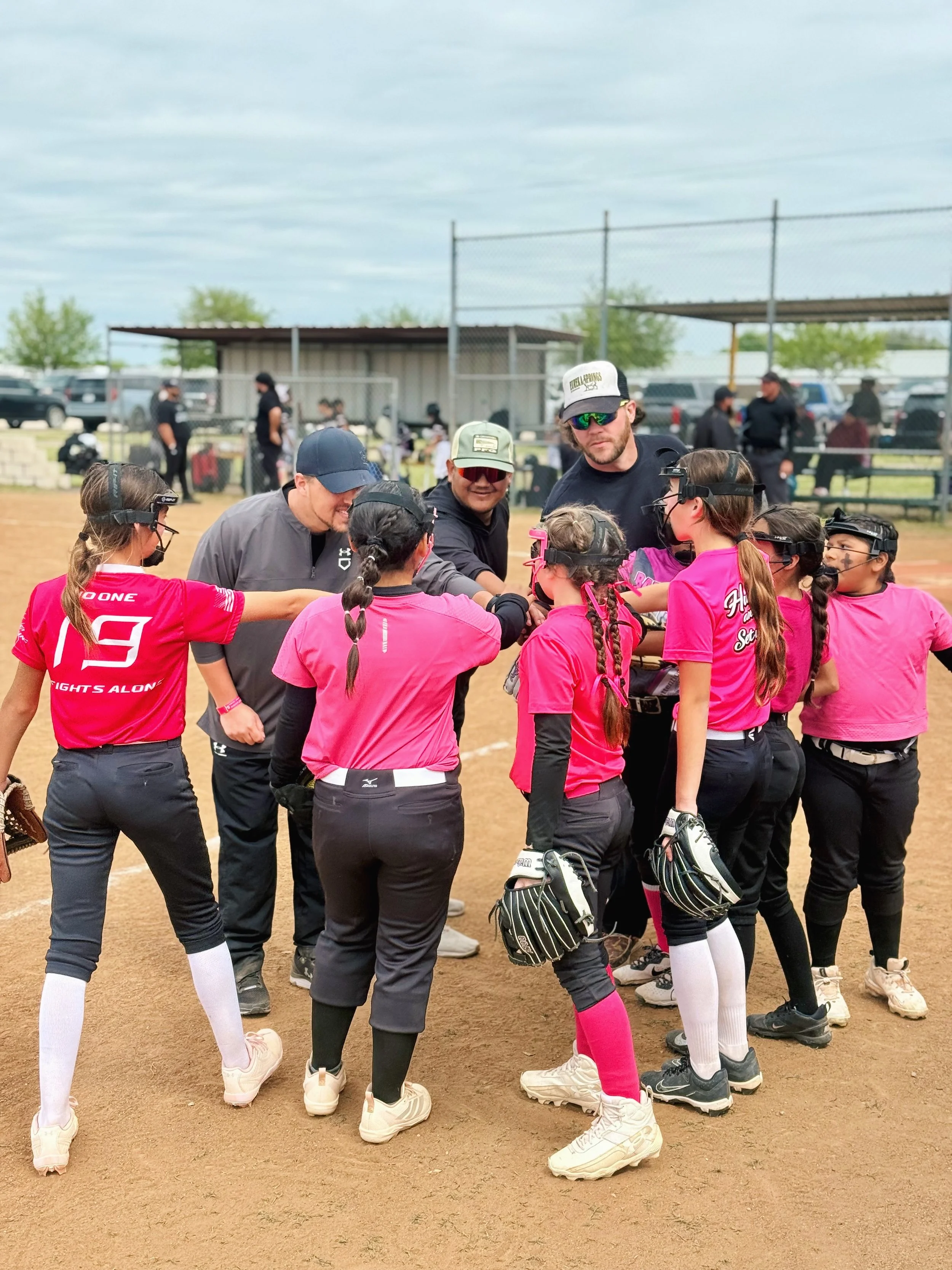 A girls' softball team in pink jerseys and black pants in a huddle on the field, with coaches giving instructions during a game.