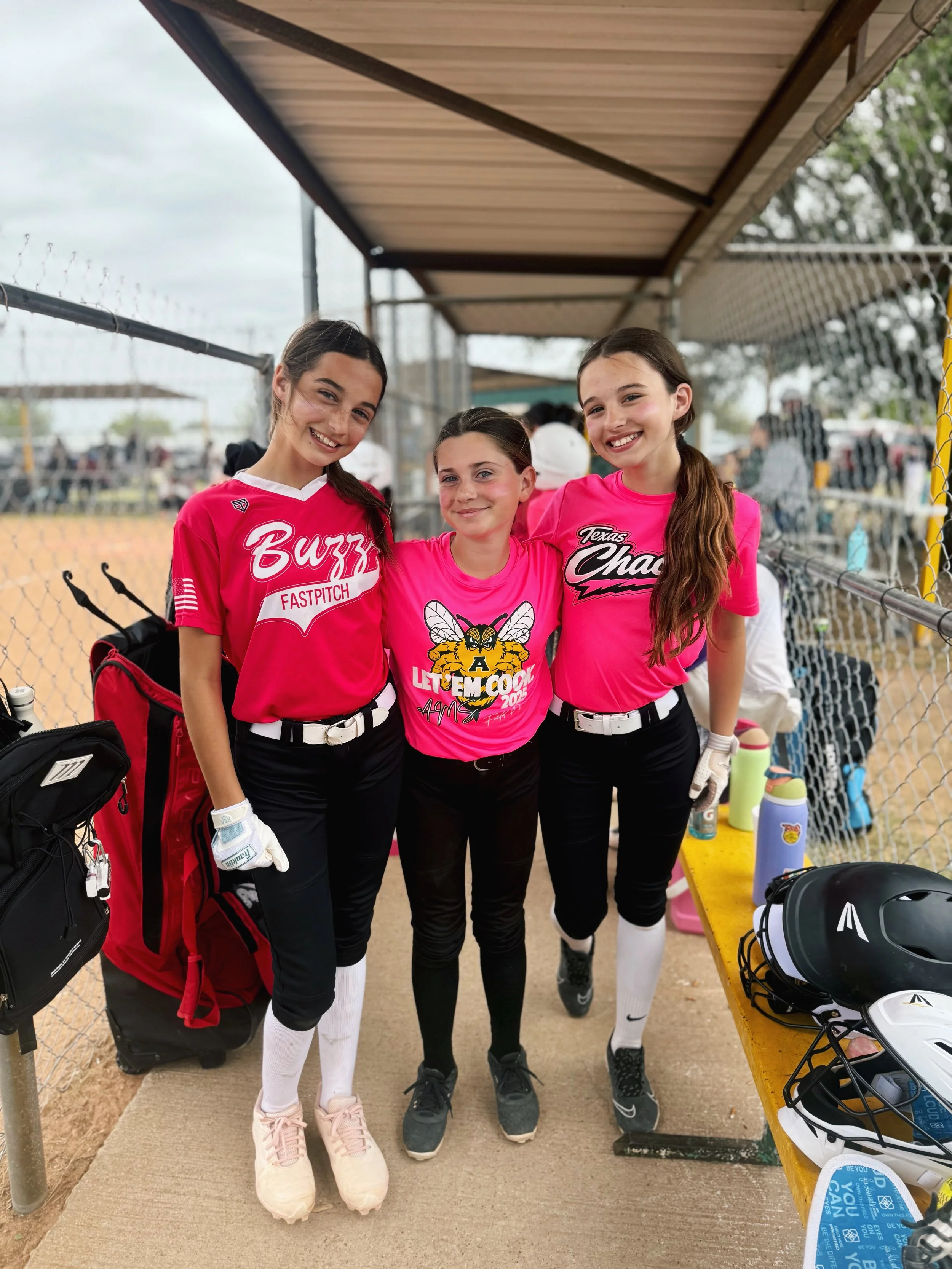 Three young female softball players in pink jerseys and black pants, posing together on a softball field, smiling, with a dugout and chain-link fence in the background.