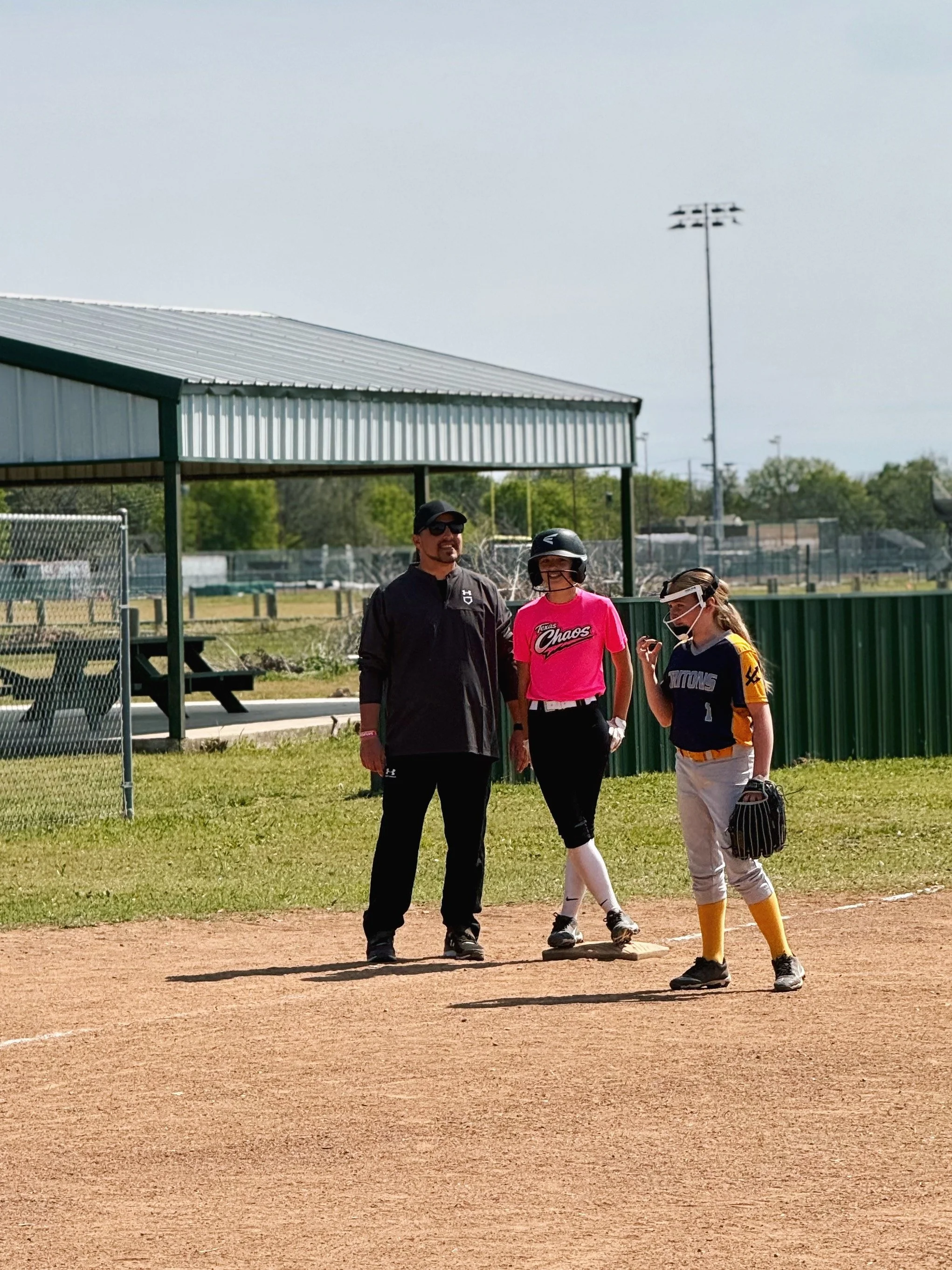 Three people on a softball field, two women and a man, standing near a base, with one woman wearing a pink shirt, another in a blue and yellow uniform, and the man in black, all wearing protective gear and sunglasses.