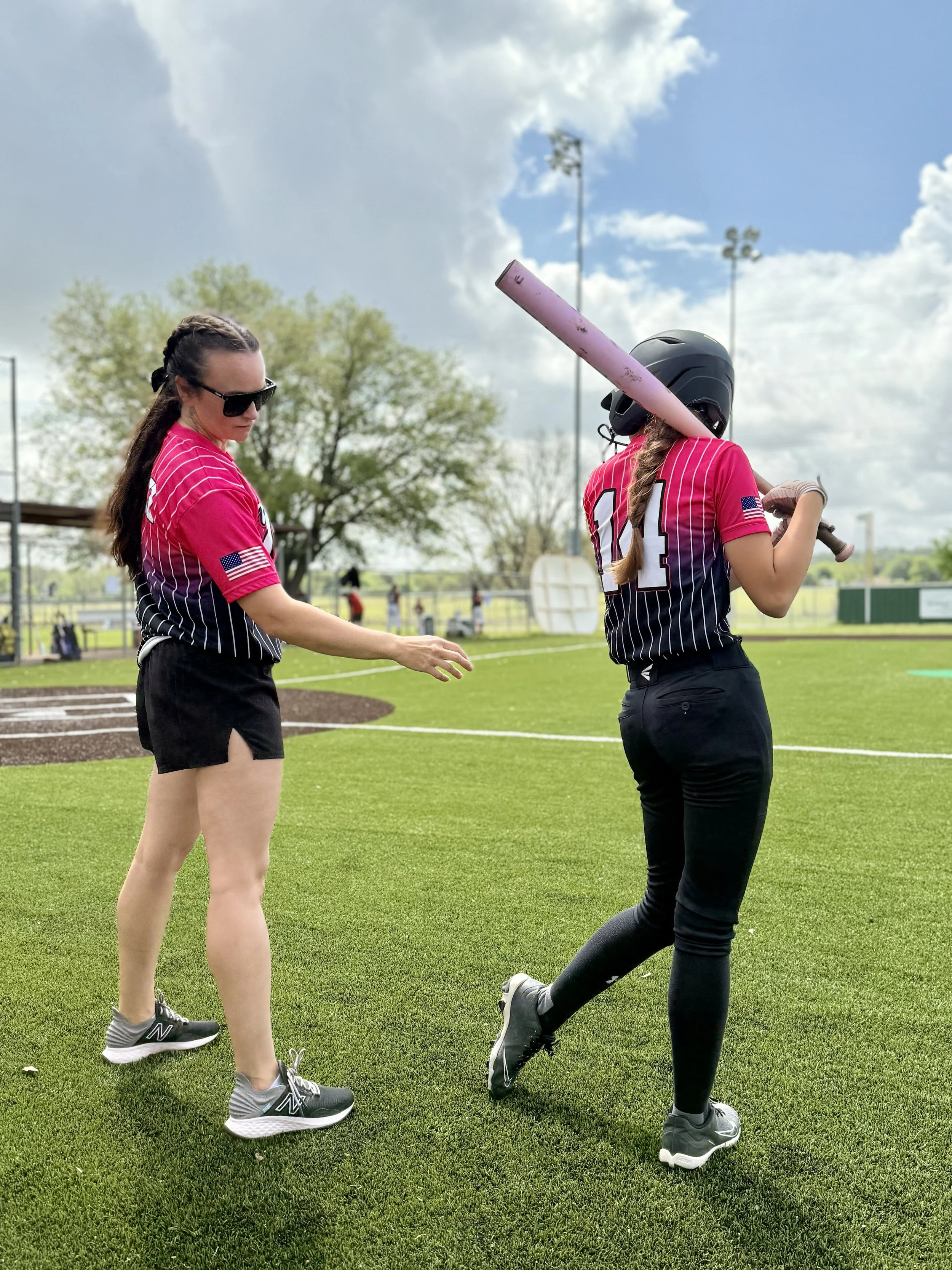 Two women in baseball uniforms with pink and black jerseys are on a baseball field. One woman is holding a bat and wearing a helmet, while the other is approaching her. The field has green grass and a baseball diamond, with a few trees and a cloudy s