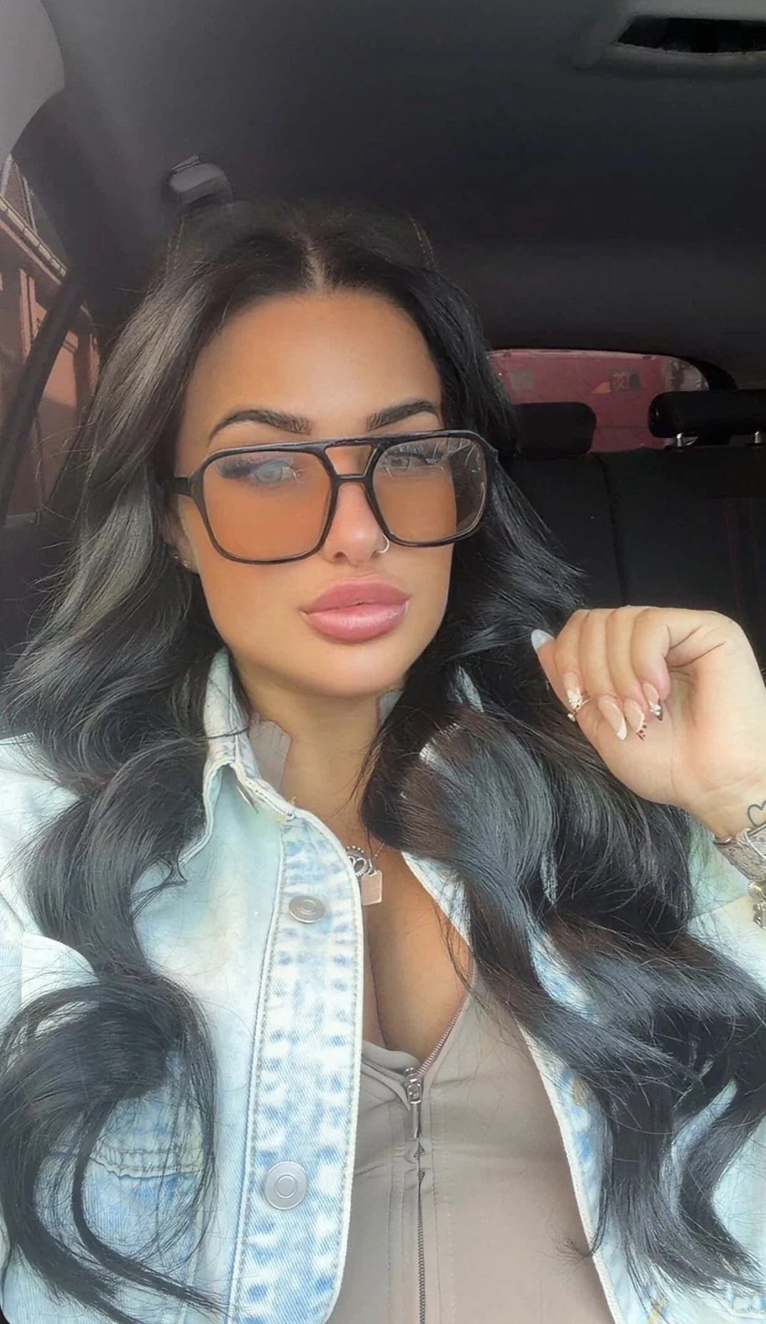 A woman with long, dark wavy hair wearing large orange-tinted glasses, a light-colored denim jacket, and a nose ring, taking a selfie inside a vehicle.