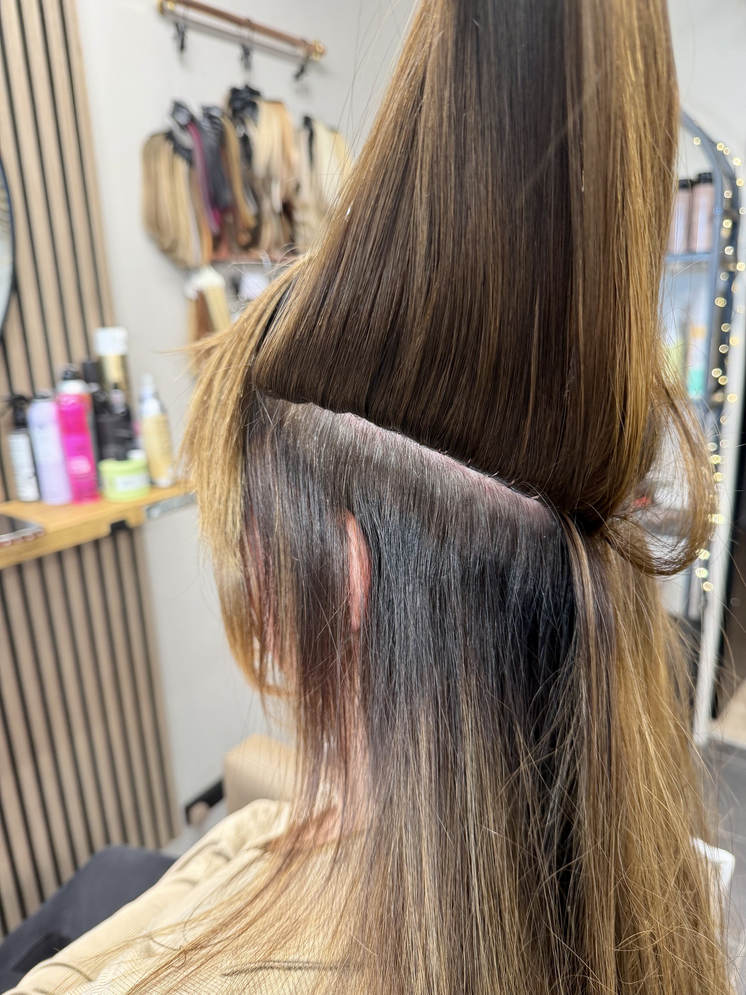 Close-up of a person's hair after a professional hair coloring or treatment, with smooth, shiny, straight, and blended highlights and lowlights in shades of brown and blonde, set in a salon with shelves of hair products in the background.