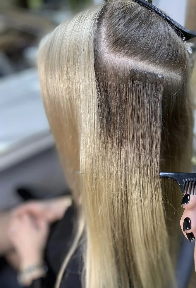 Close-up of a stylist cutting and coloring a woman's long, straight blonde hair, with sections of dark and light shades visible.