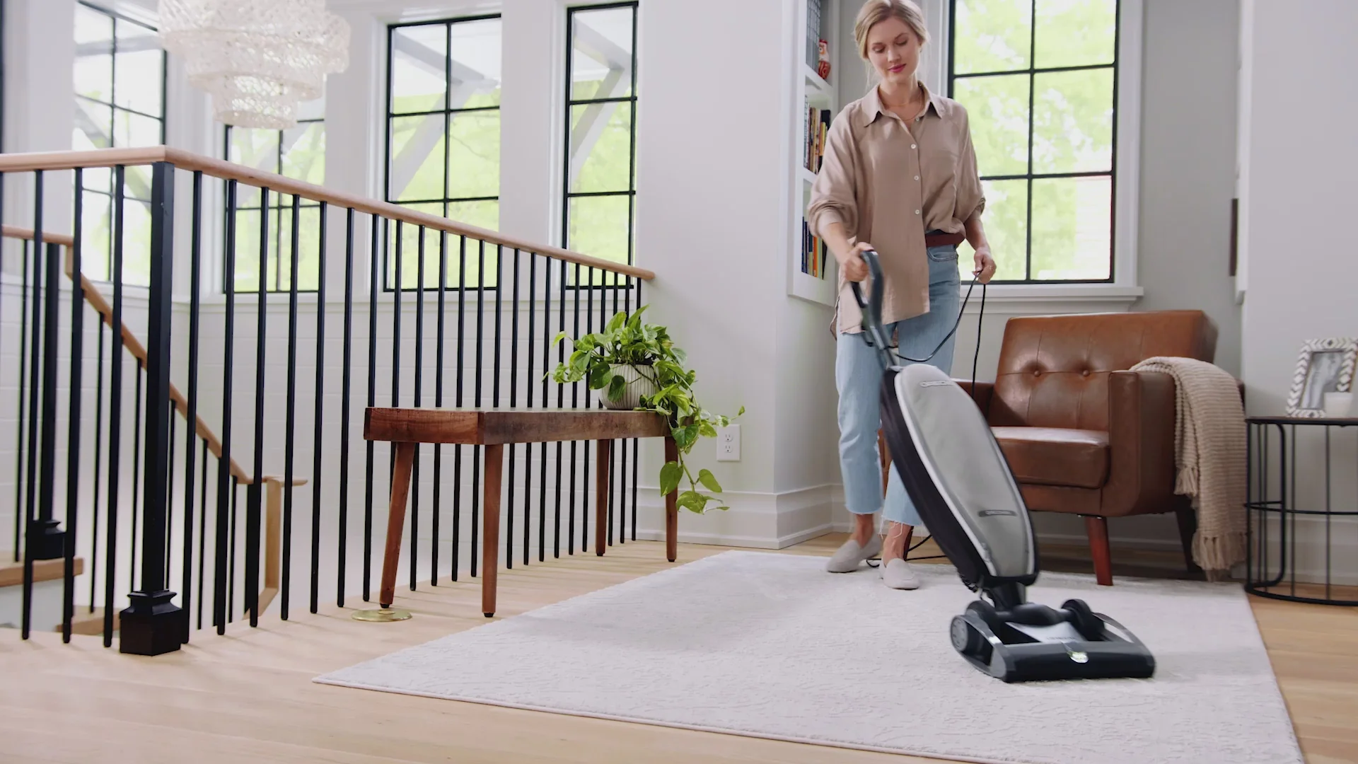 A woman in casual clothing vacuuming a living room with large windows and a brown sofa.