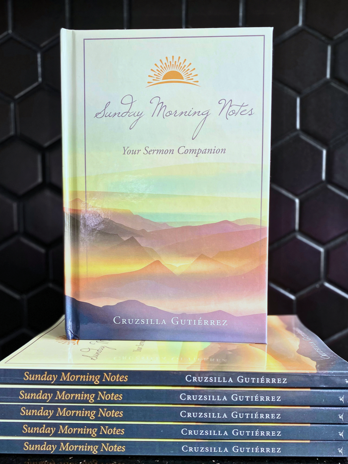 A paperback book titled "Sunday Morning Notes" by Cruzsilla Gutiérrez with a pastel-colored mountain landscape on the cover, standing upright in front of several copies stacked horizontally.