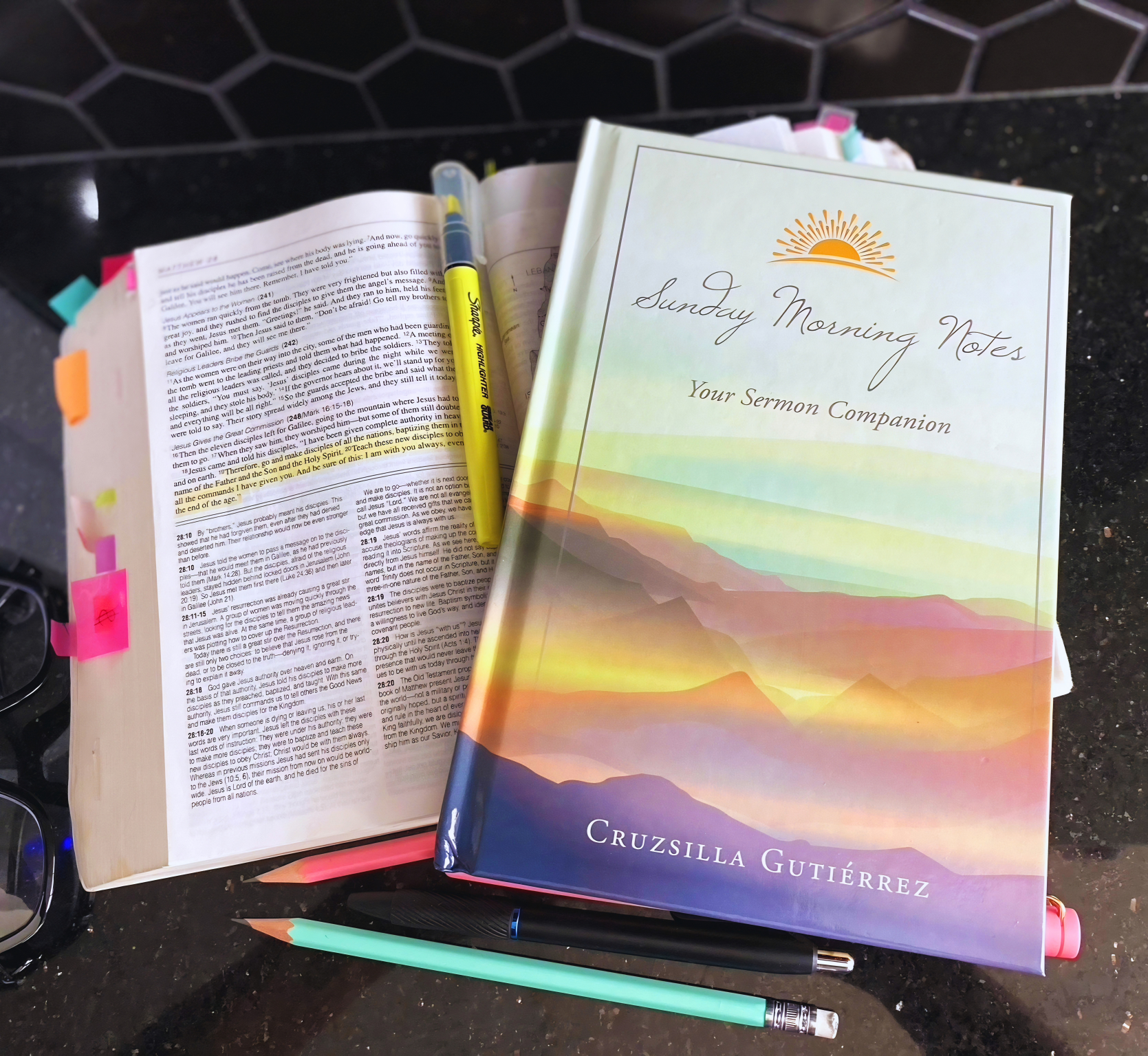 A closed book titled 'Sunday Morning Notes: Your Sermon Companion' by Cruzsilla Gutiérrez is on a black countertop, with an open Bible next to it, along with a yellow Sharpie marker, a black pen, a mint green pen, and a pink highlighter.