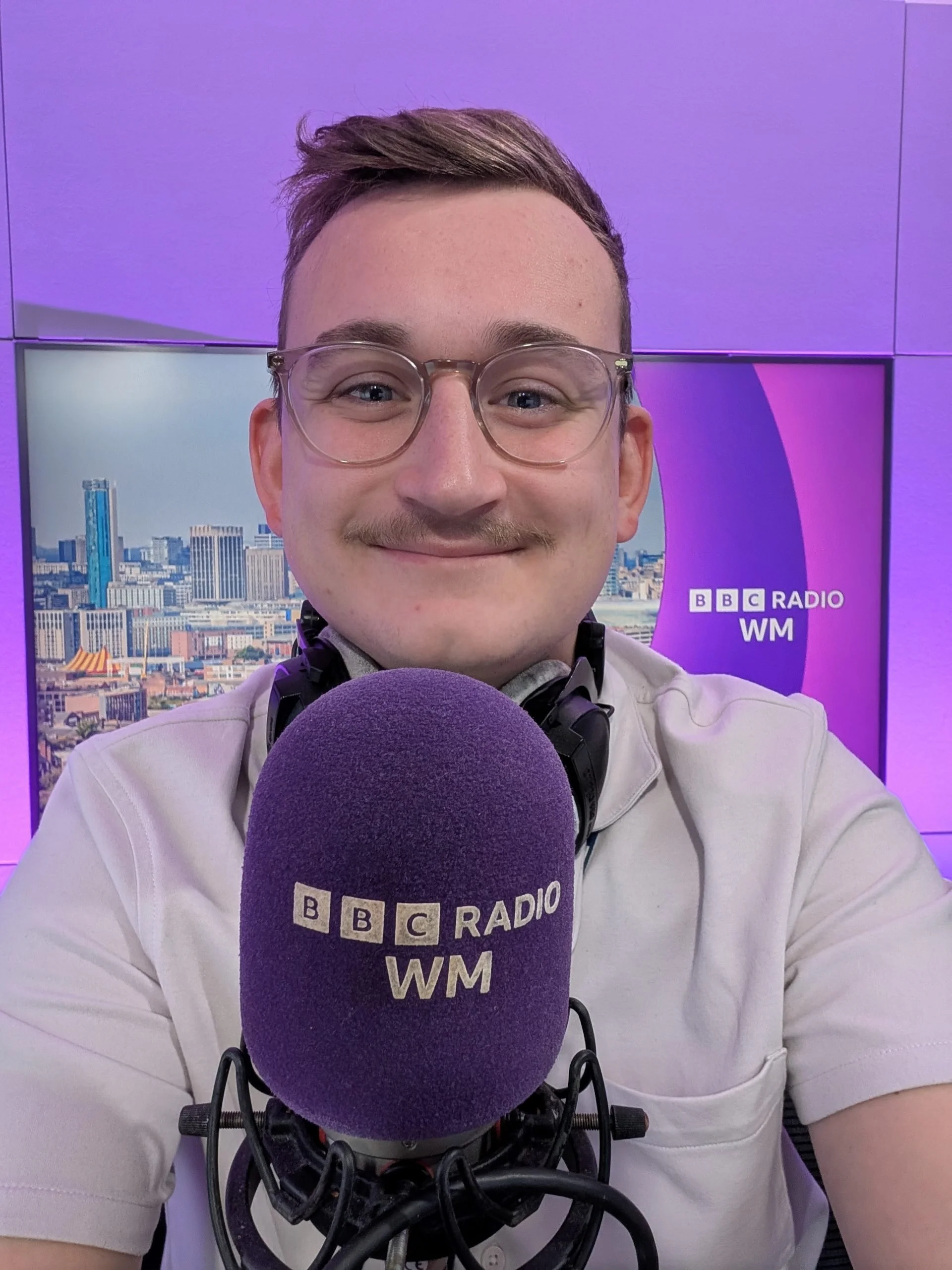 A young man with glasses and a smile taking a selfie in a radio studio. He is wearing a white shirt, with a purple foam microphone cover labeled 'BBC Radio WM' in front of him, and over-ear headphones around his neck. The background features a purple and blue cityscape with the BBC Radio WM logo.