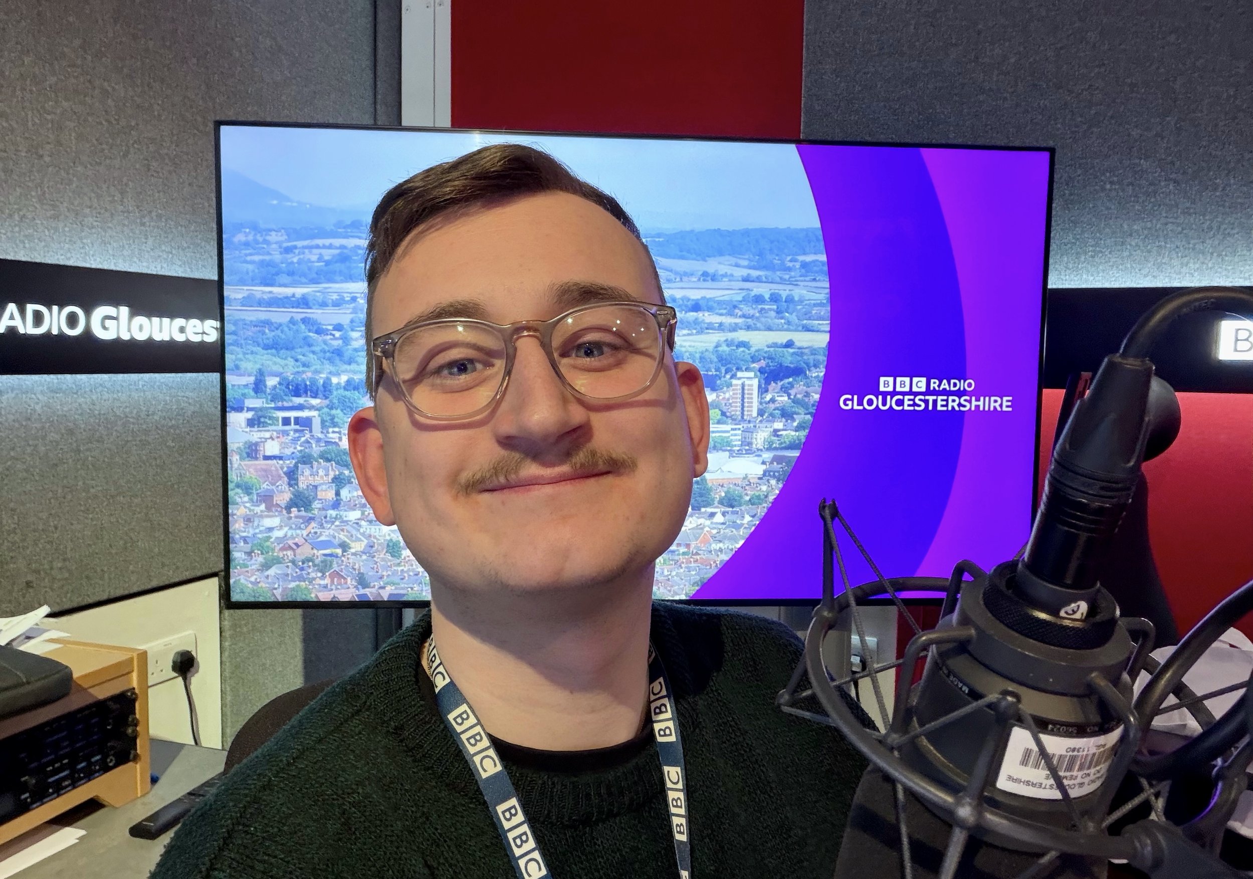 Filip with glasses smiling in a radio studio with a microphone and BBC Gloucestershire signage and background.