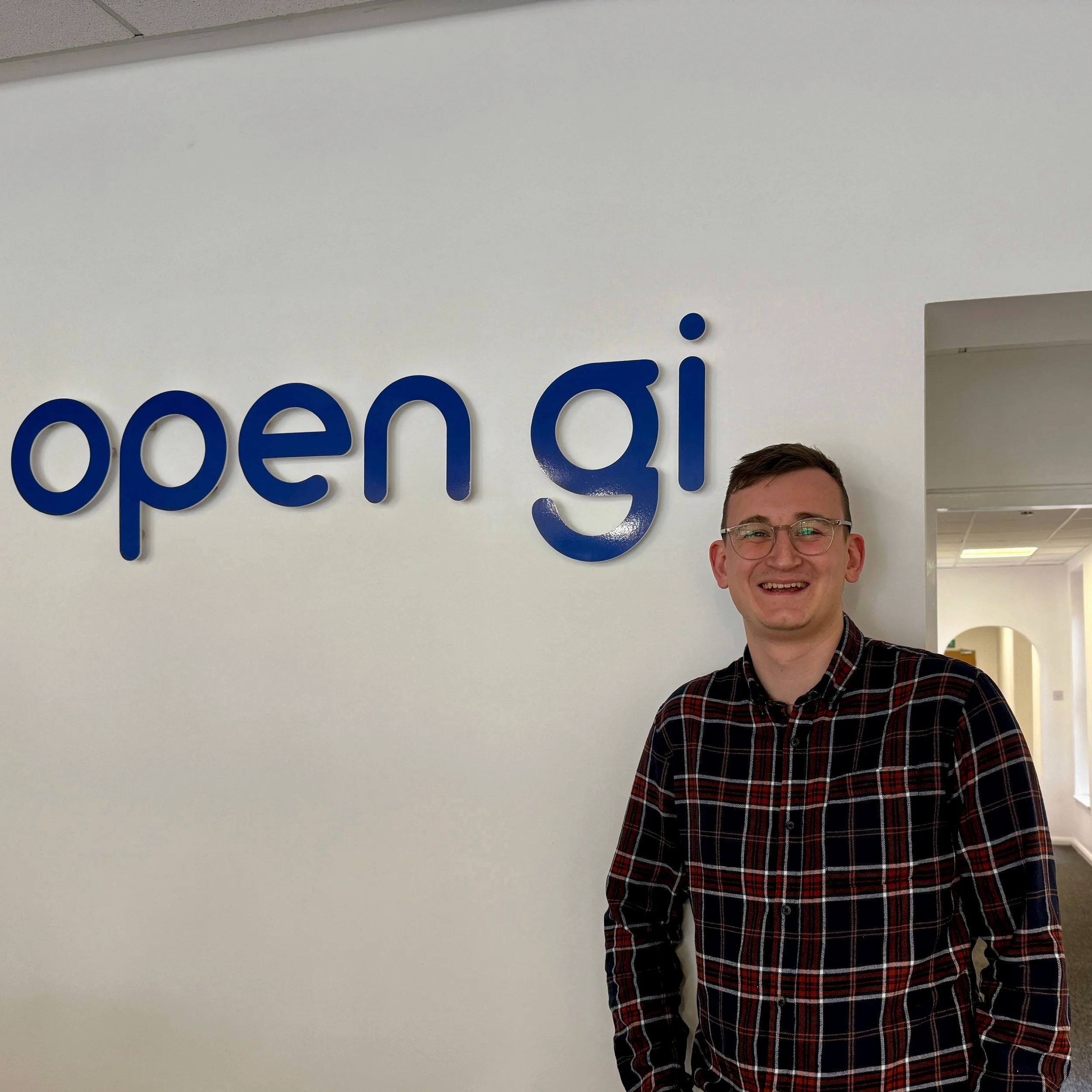 Filip with glasses and a plaid shirt smiling while standing next to a wall with the Open GI logo.