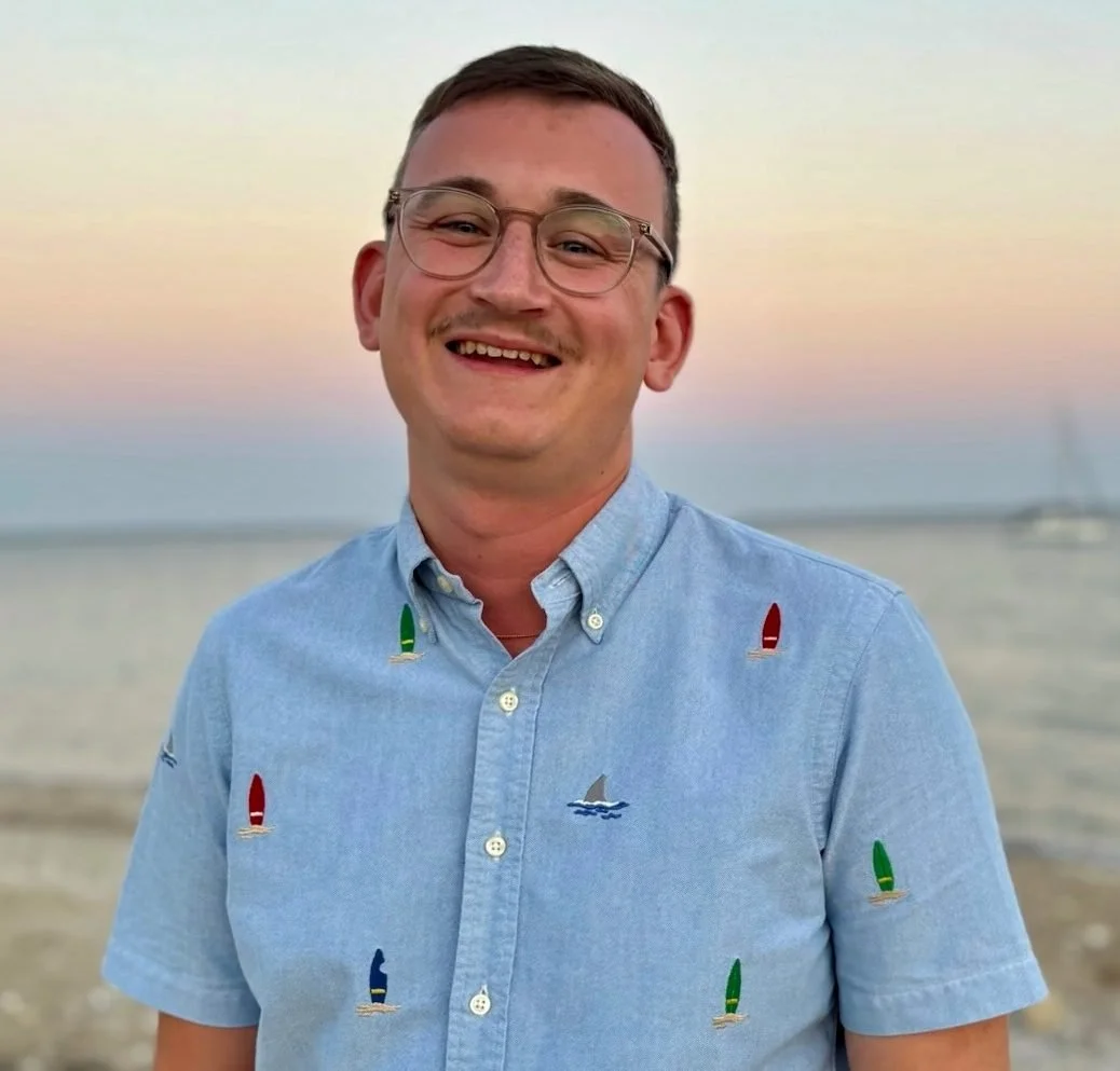 A smiling young man with glasses, wearing a light blue shirt with sailboat embroidery, standing outdoors at sunset or dusk by the water.