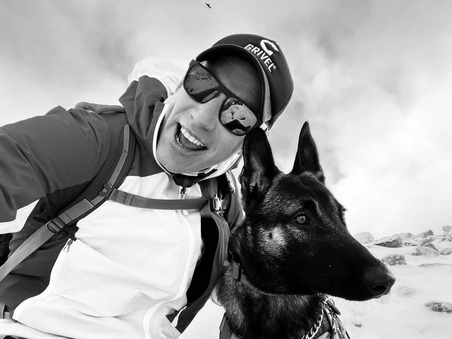 A man wearing sunglasses, a helmet, and outdoor gear taking a selfie with a black and brown dog on a rocky, snowy landscape.