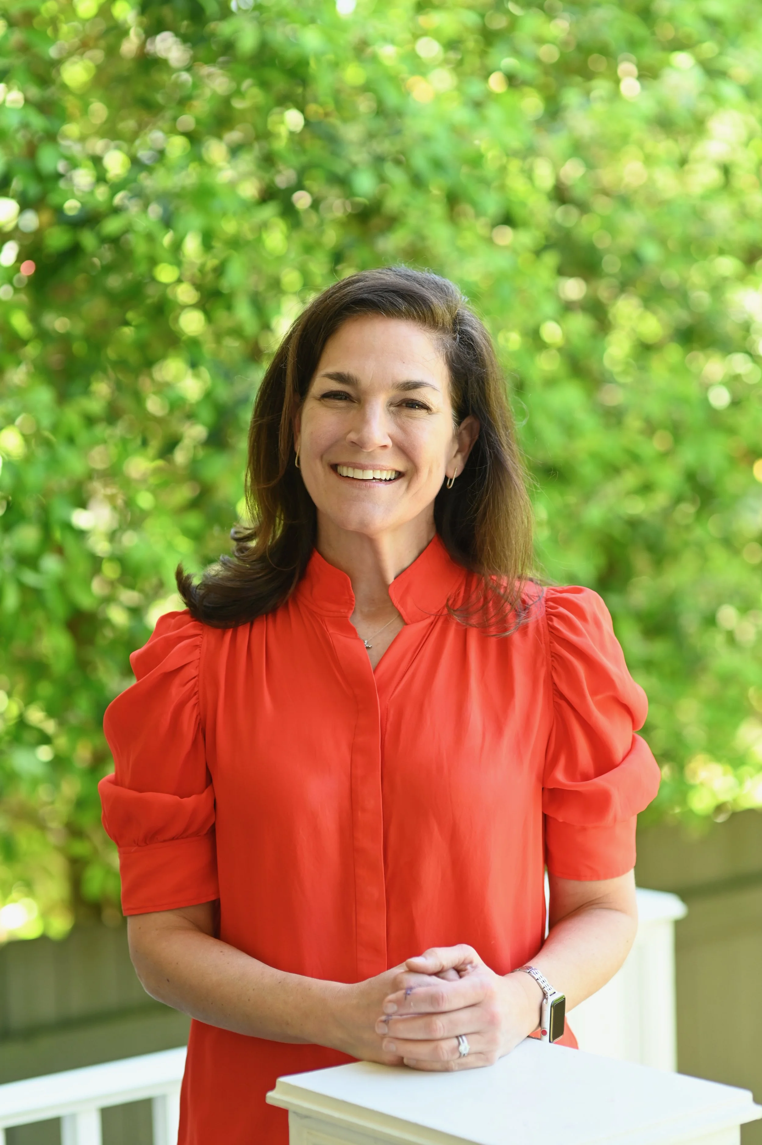 A smiling woman with shoulder-length brown hair, wearing a red blouse with puffed sleeves, stands outdoors with a background of green trees and sunlight filtering through the leaves.