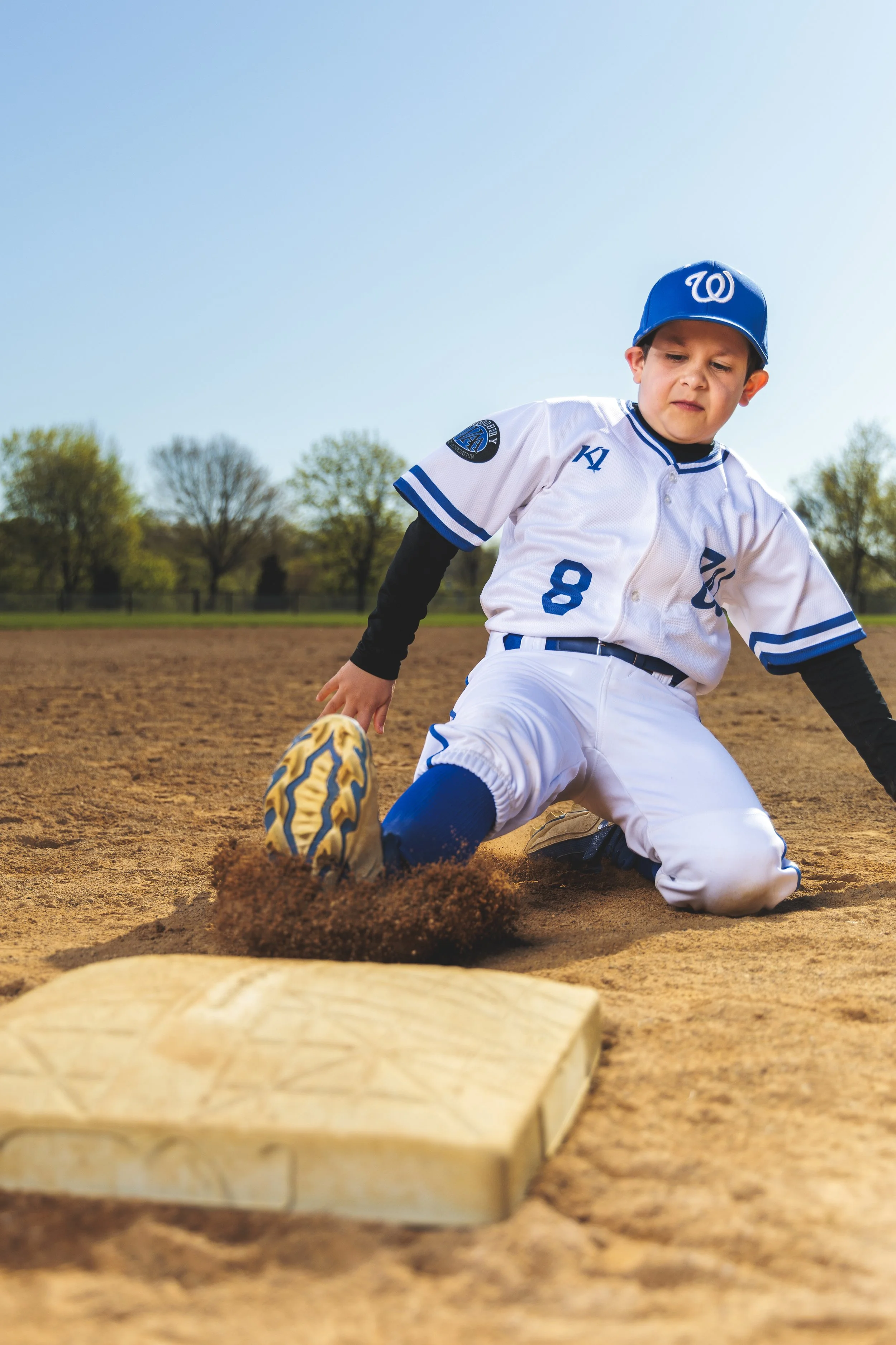 A young baseball player in a white uniform sliding into a base on a dirt field, with trees and a clear blue sky in the background.