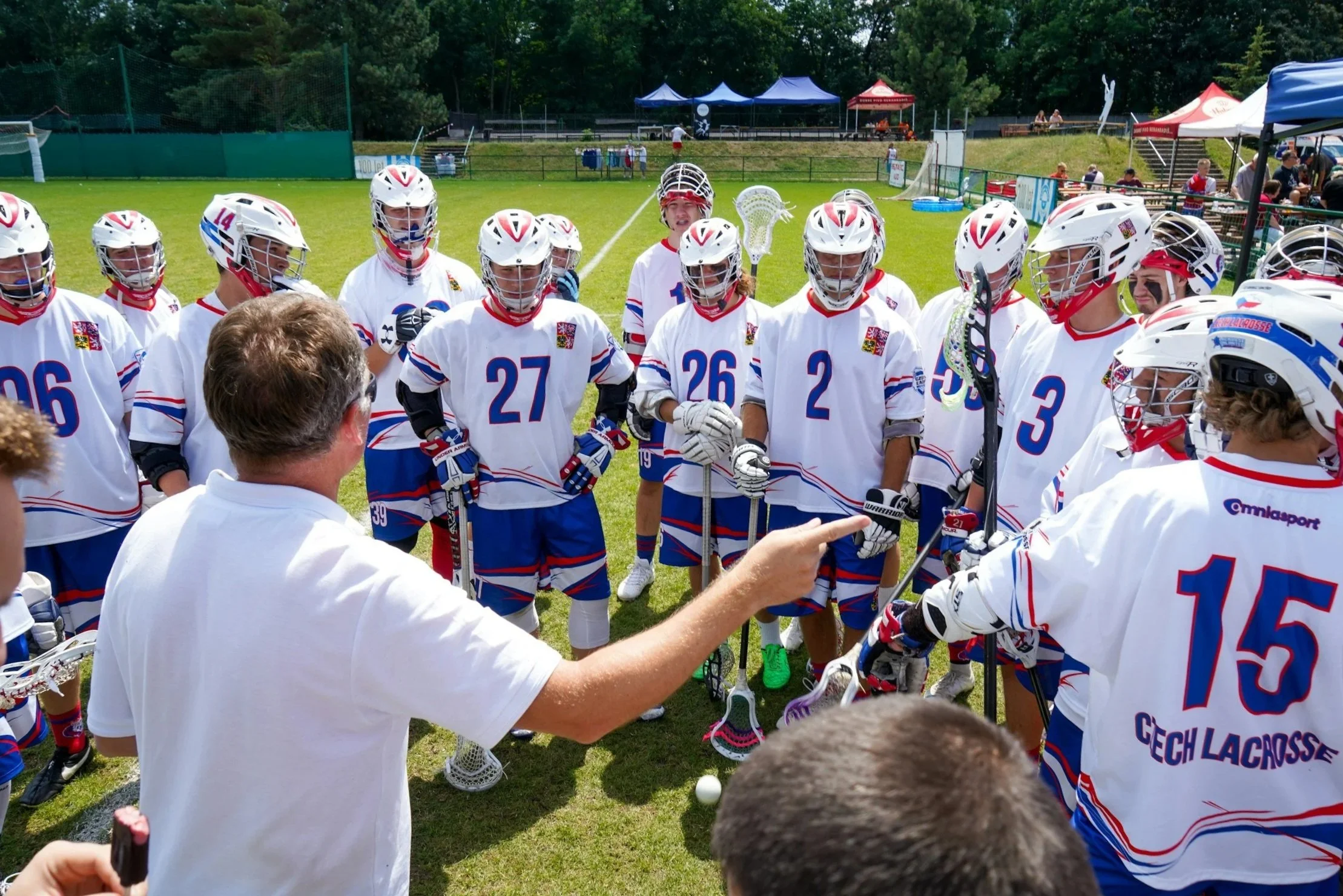 A group of lacrosse players in white jerseys with red and blue accents, wearing helmets and holding lacrosse sticks, gathered during a game or practice on a grassy field with tents and spectators in the background.