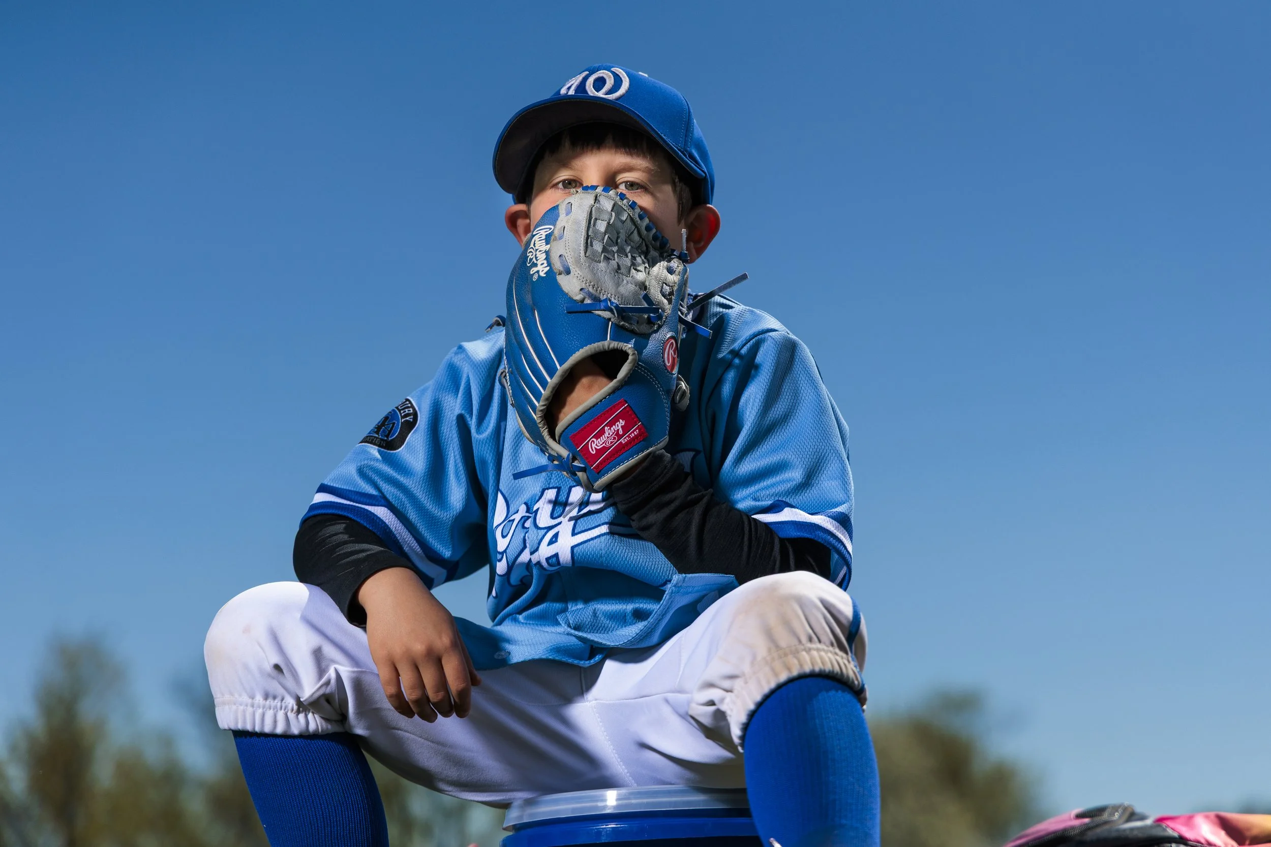 Young boy wearing a blue baseball cap, blue jersey, and white pants, sitting on an overturned bucket outdoors, holding a baseball glove to his face against a clear blue sky.
