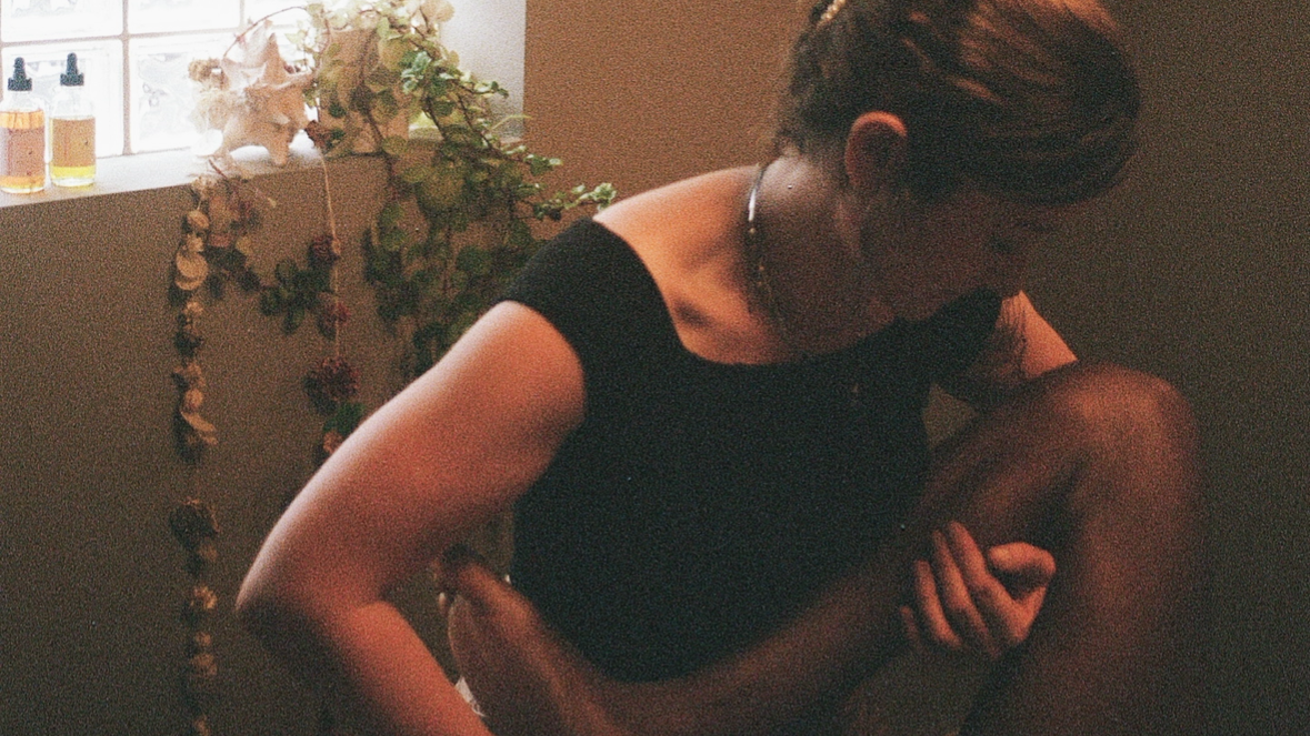 Simone Crowe, Licensed Massage Therapist, wearing a black top, performs Thai massage leg stretches for her client, near a window with bottles and dried flowers.