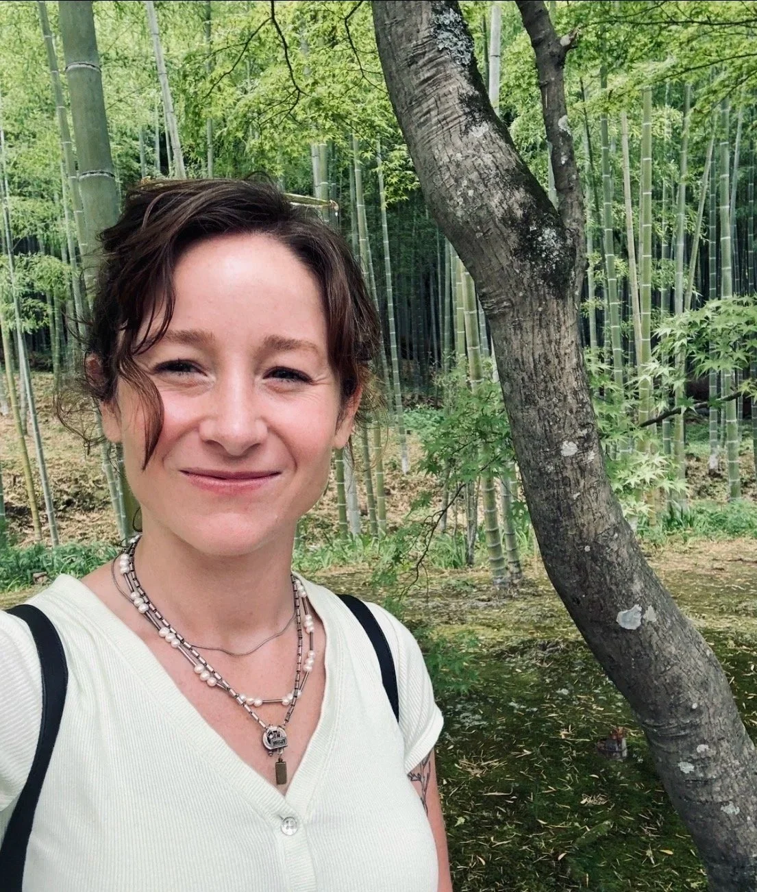 Simone Crowe, massage therapist, smiling in a lush green bamboo forest, wearing a light-colored shirt and layered necklaces.