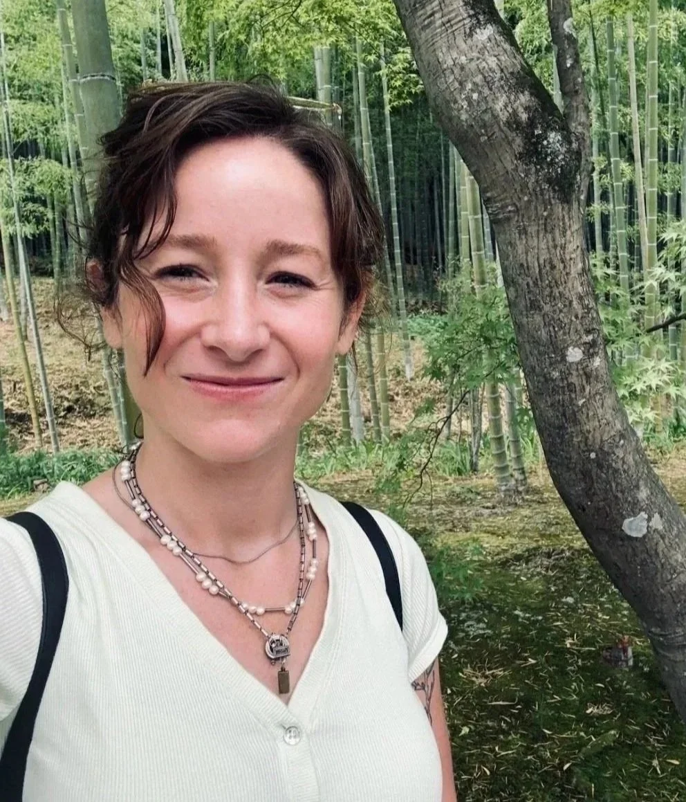 A woman with short brown hair smiling outdoors in a forest with bamboo and trees in the background.