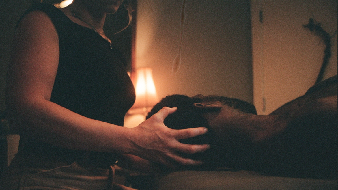 Simone Crowe, Licensed Massage Therapist gently holds a man's head while he lies on a massage table in a dimly lit room.