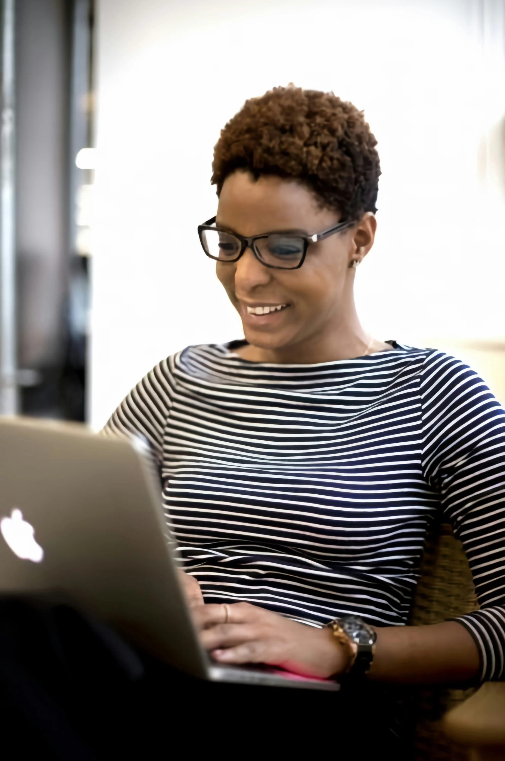 Woman with short curly hair, glasses, and a striped shirt smiling while using a laptop.