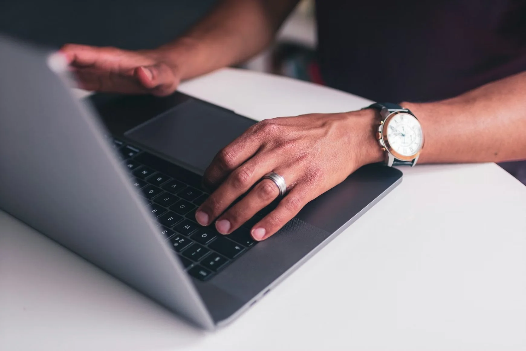 Close-up of a person typing on a laptop keyboard, wearing a wristwatch and a ring on their left hand.