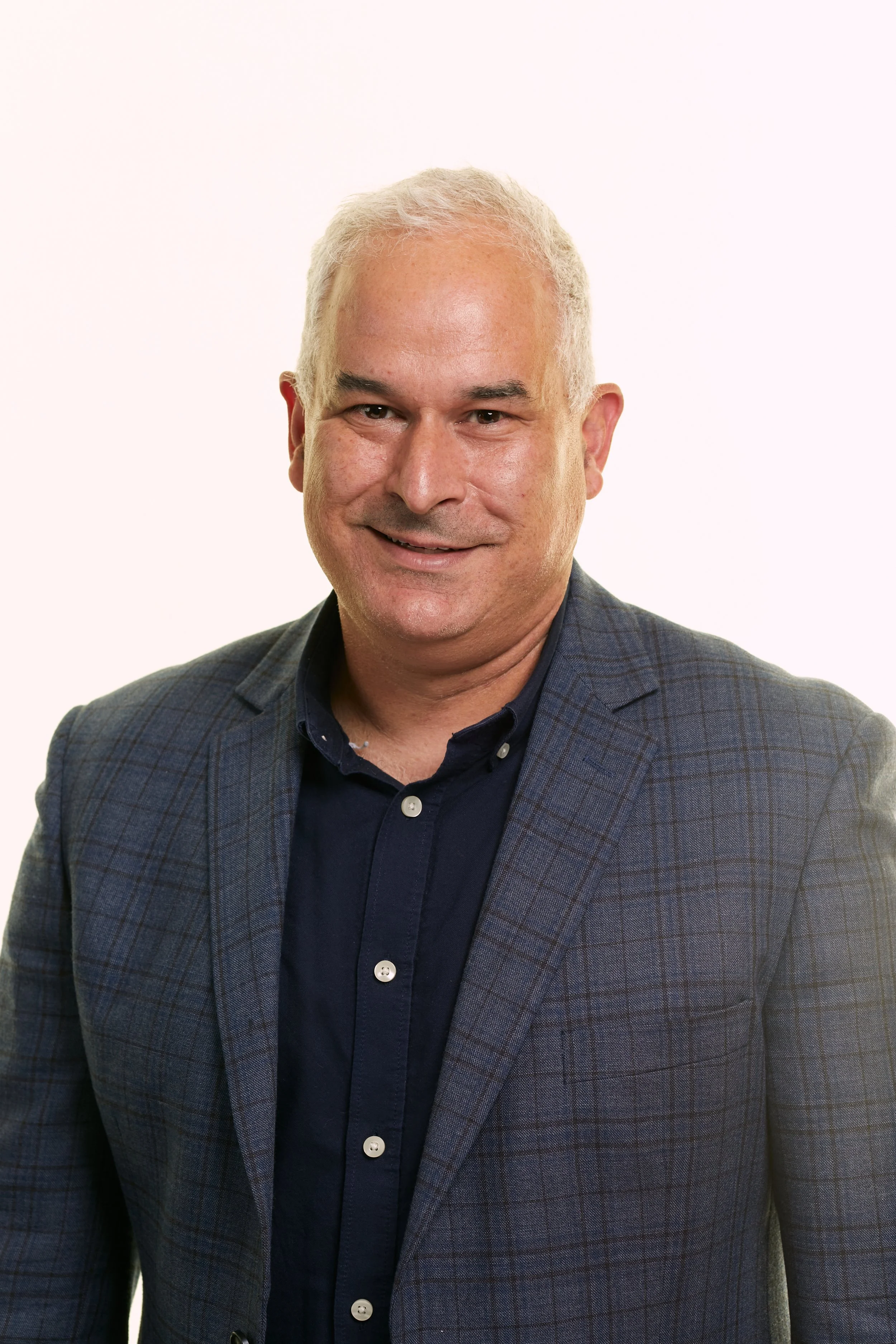 Headshot of a middle-aged man with short gray hair, wearing a dark navy shirt and a gray plaid blazer, smiling against a plain white background.