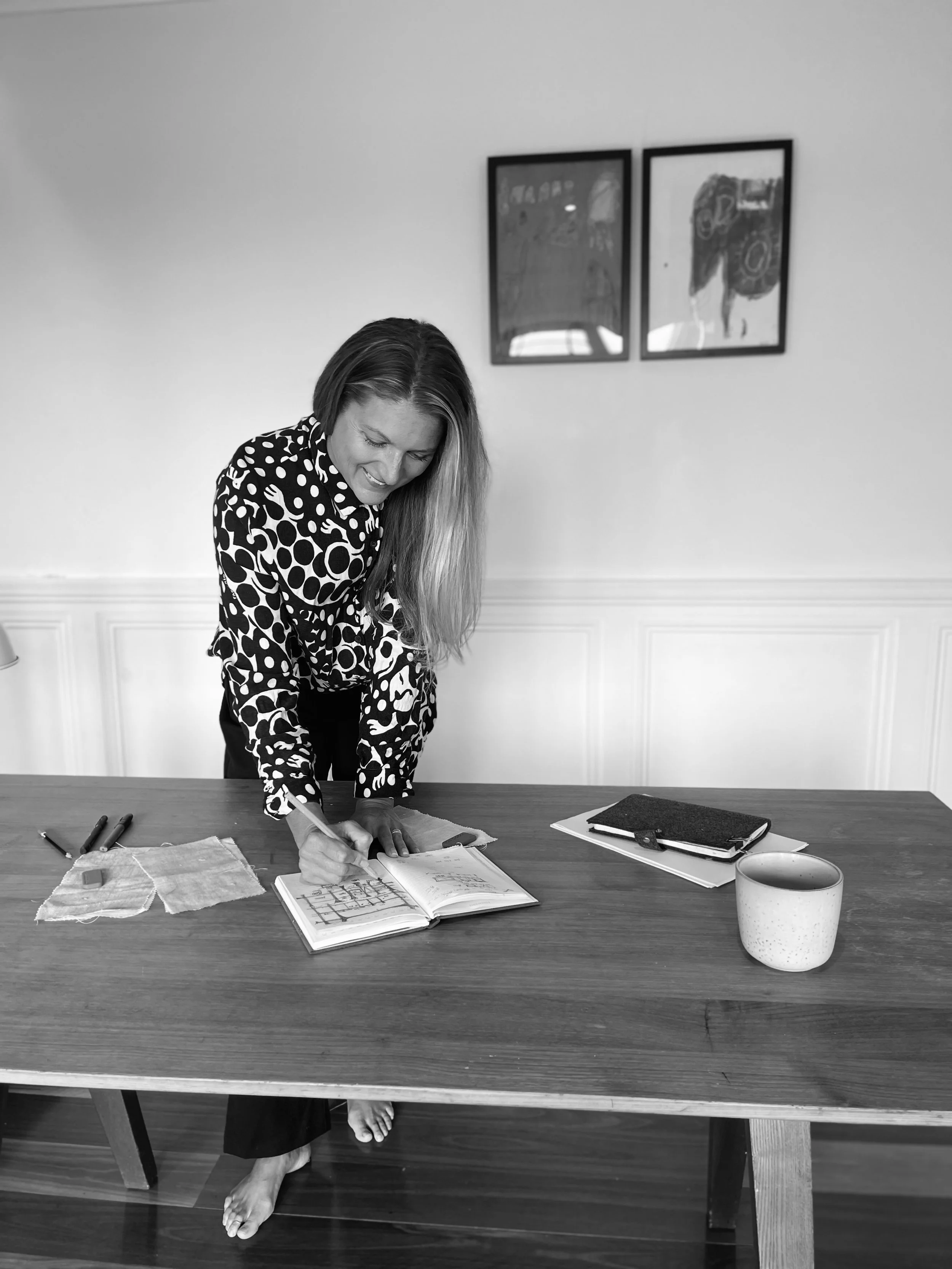 A woman with long hair, wearing a polka dot shirt and a skirt, is standing at a wooden table. She is leaning over and drawing in a sketchbook. There are pens, a fabric swatch, and notebooks on the table. The woman is barefoot, and there are two framed pictures on the plain wall behind her.
