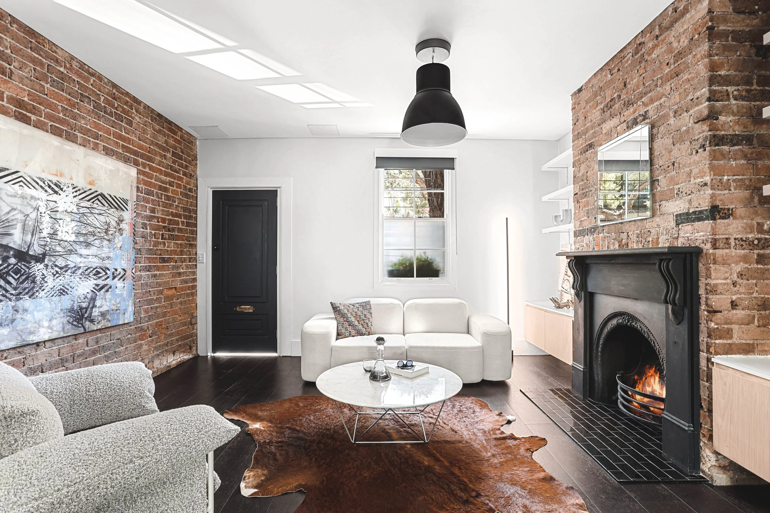 A living room with exposed brick walls, a white sofa, a black fireplace with a fire, a cowhide rug, black and gray furniture, and a window with a view of trees outside.