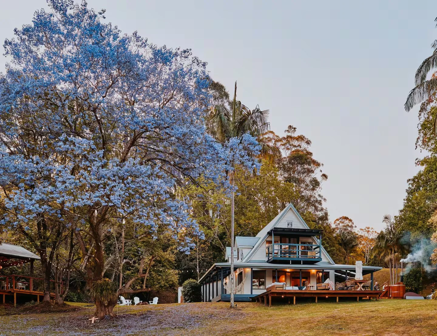 A cozy white house with a large porch and outdoor seating, surrounded by tall trees, including one with blue blossoms, during sunset in a rural setting.