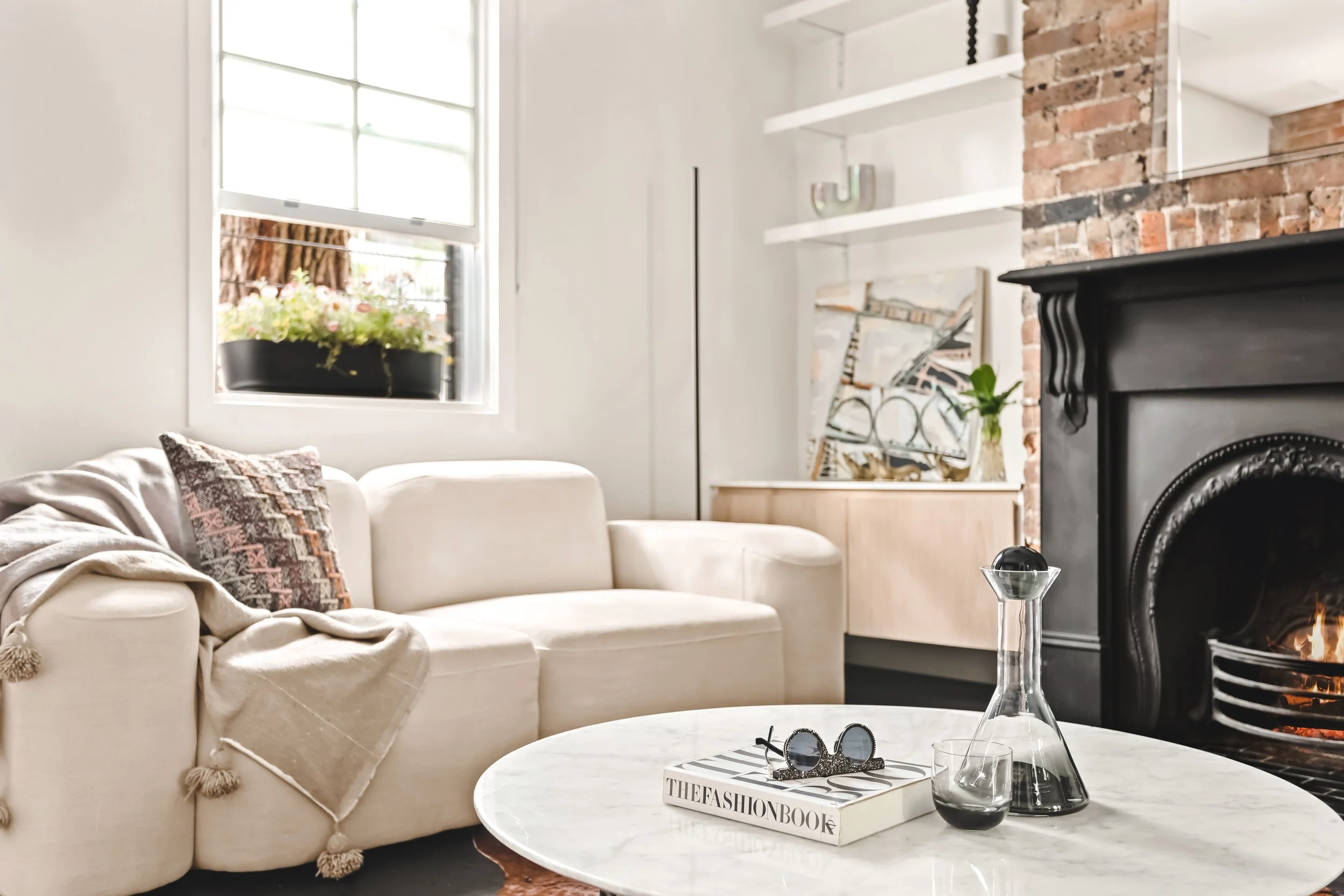 A cozy living room with a white sofa decorated with colorful throw pillows, a round marble coffee table with sunglasses and a fashion book, a black fireplace with a glass of whiskey and a glass sculpture, and a window with a flower box. There are white shelves and a brick wall in the background.