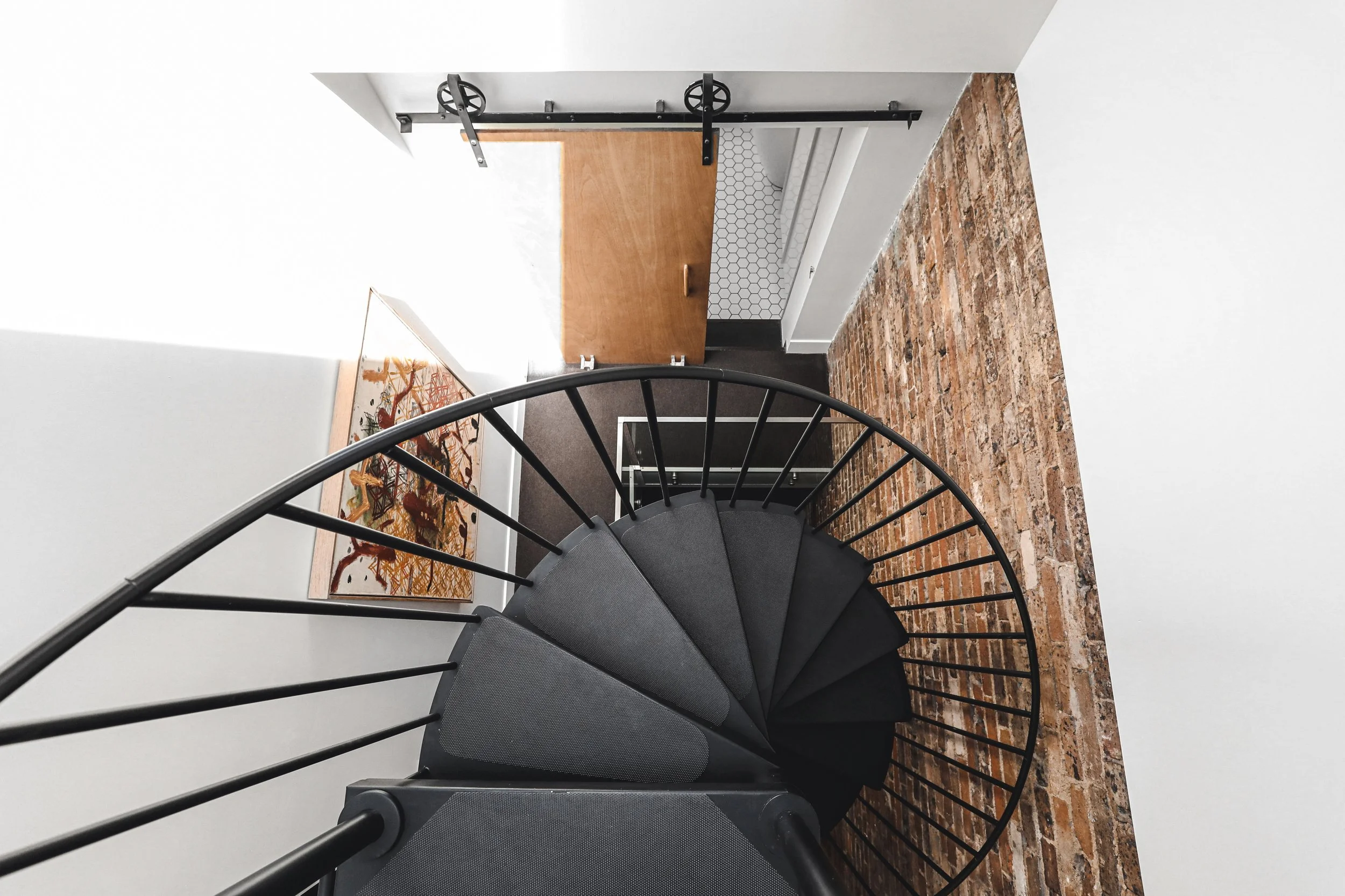 A spiral staircase with black railings and gray steps, viewed from above, leading down to a tiled floor with brick and white wall backgrounds.