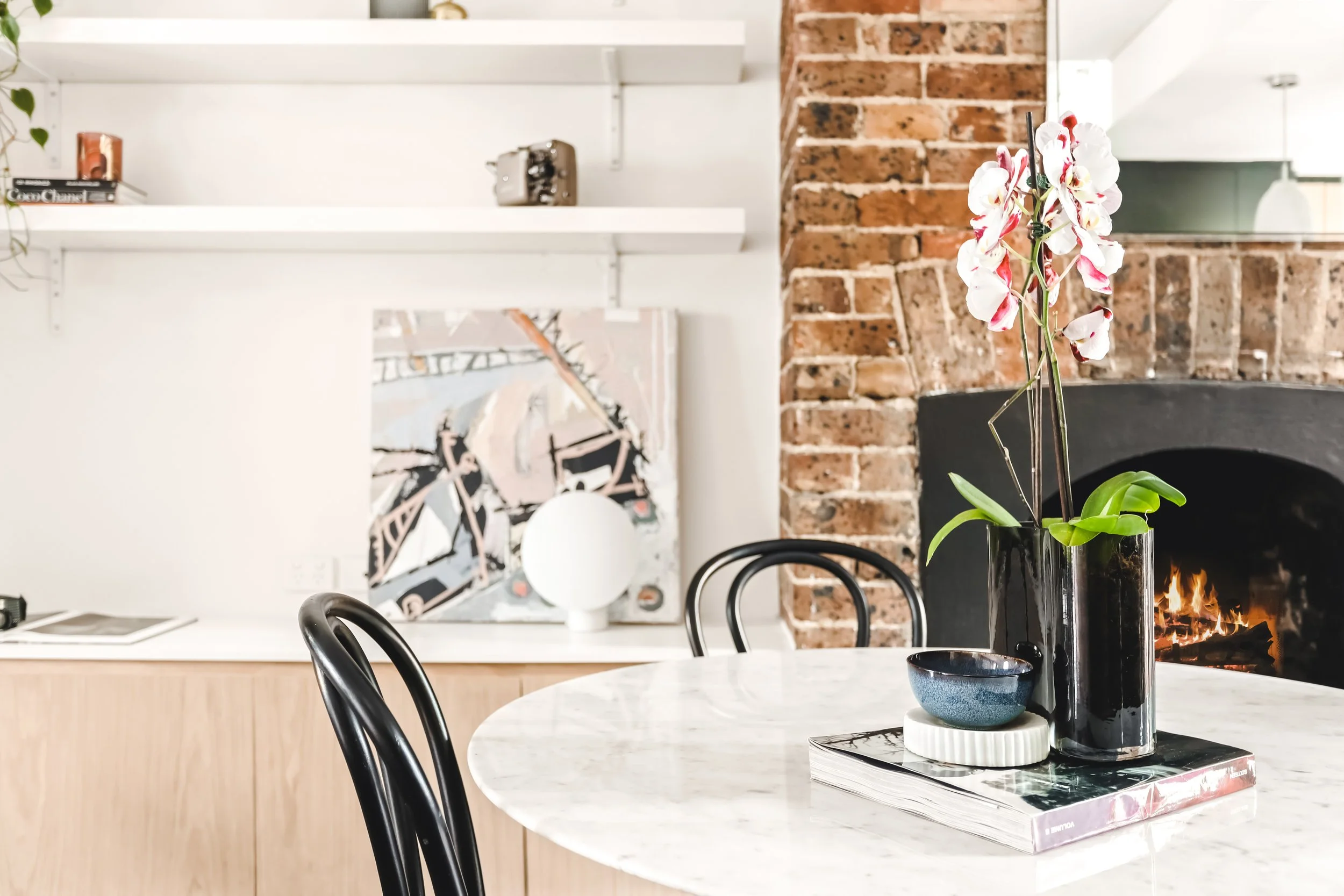 A modern kitchen or dining area with a white marble table, black chairs, a black vase with white and pink orchids, a bowl, and a stack of books on the table. The background features brushed wood cabinets, white shelves with decorative items, and an exposed red brick wall with a fireplace.