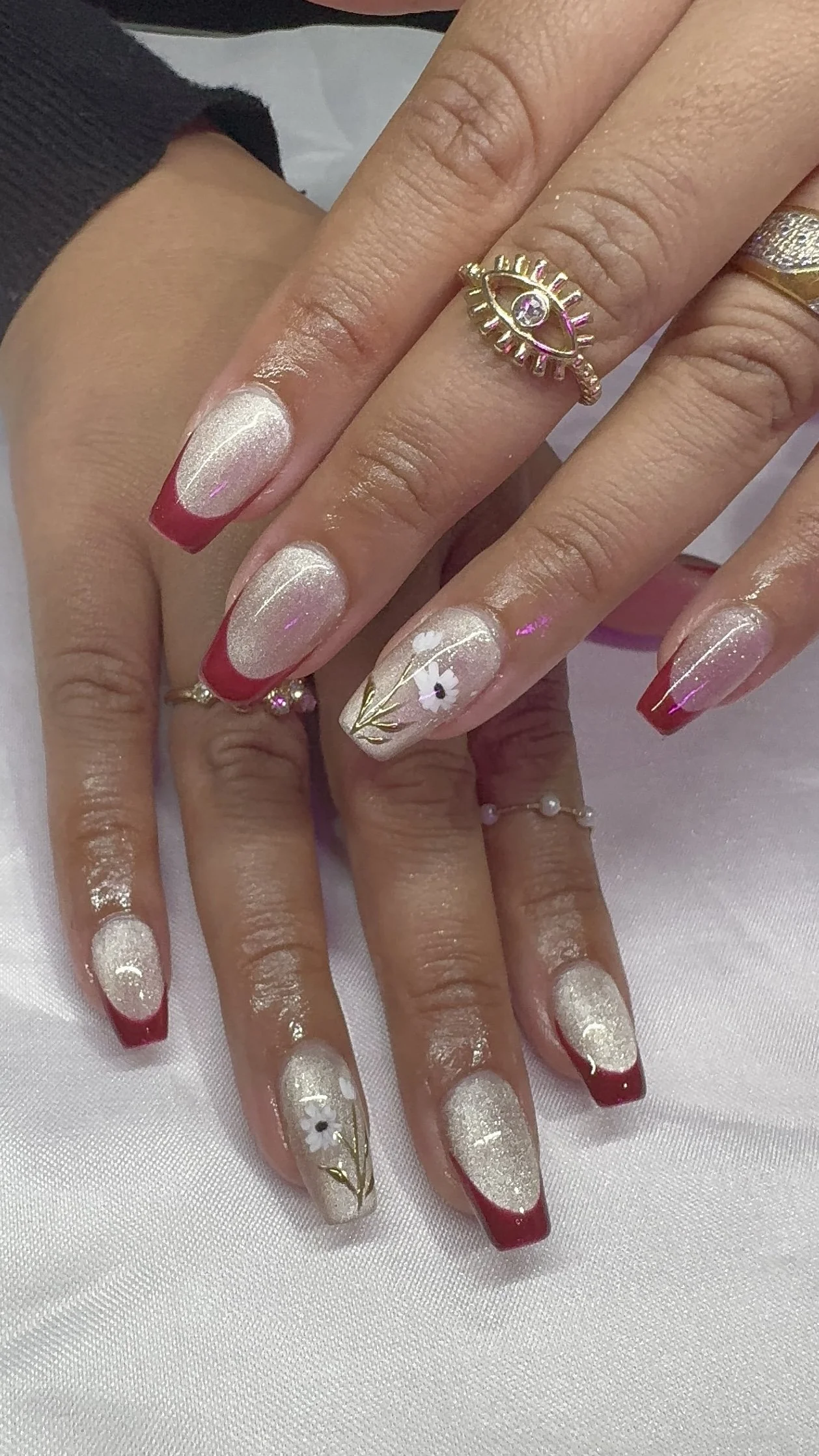 Close-up of manicured hands with long oval nails painted in a glittery white color with red tips, and one nail with floral and gold accents. The individual is wearing multiple rings, including a gold eye-shaped ring with a gem in the center and a textured silver ring. The background is a white fabric surface.