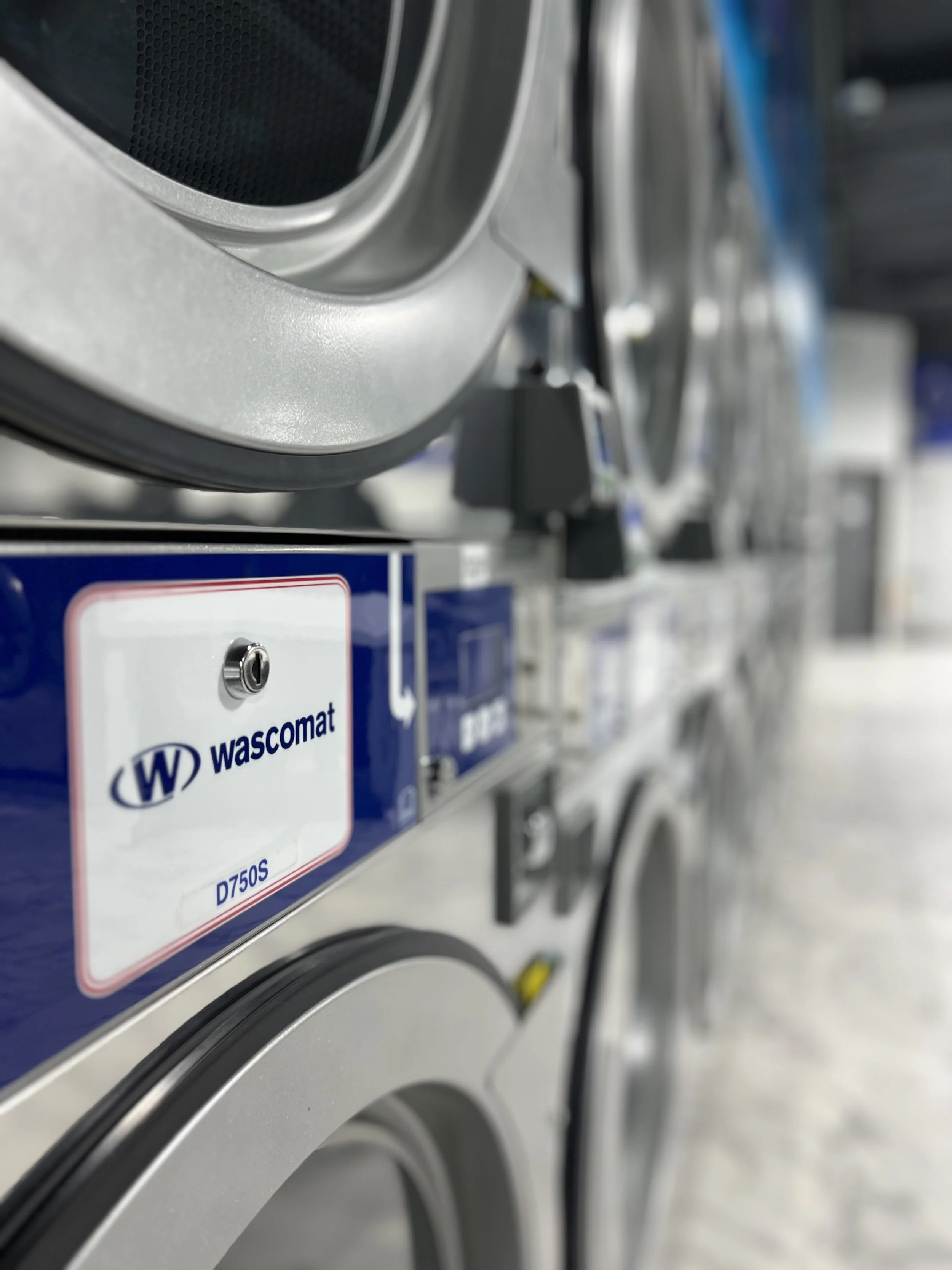 Close-up of a commercial washing machine with a blue and white label that has the brand name 'Wascomat' and model number 'D750S' seen on the front.