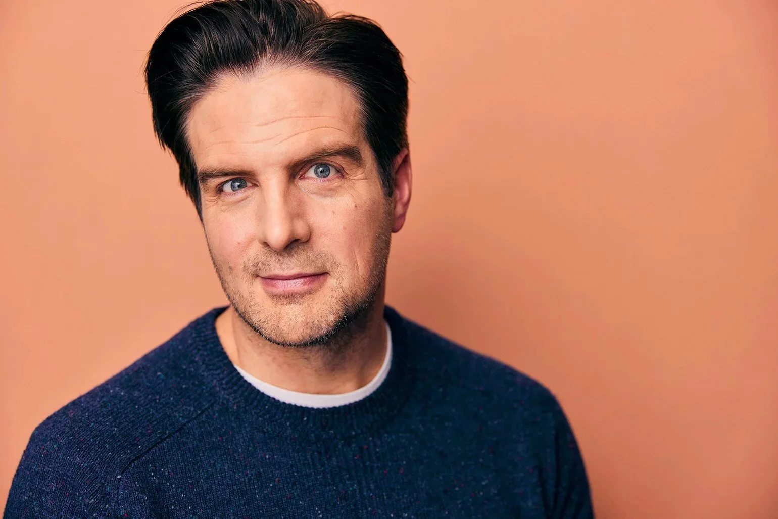 A man with dark hair, blue eyes, and light skin wearing a navy blue sweater stands against a peach-colored background.