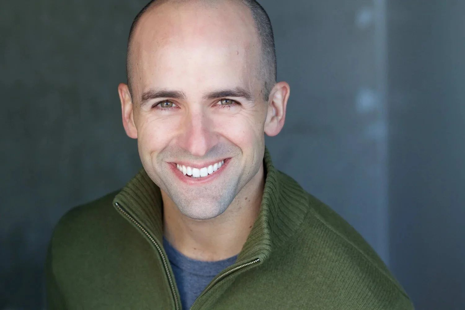 Headshot of a smiling man with short dark hair, light skin, wearing a green zip-up sweater, against a dark background.