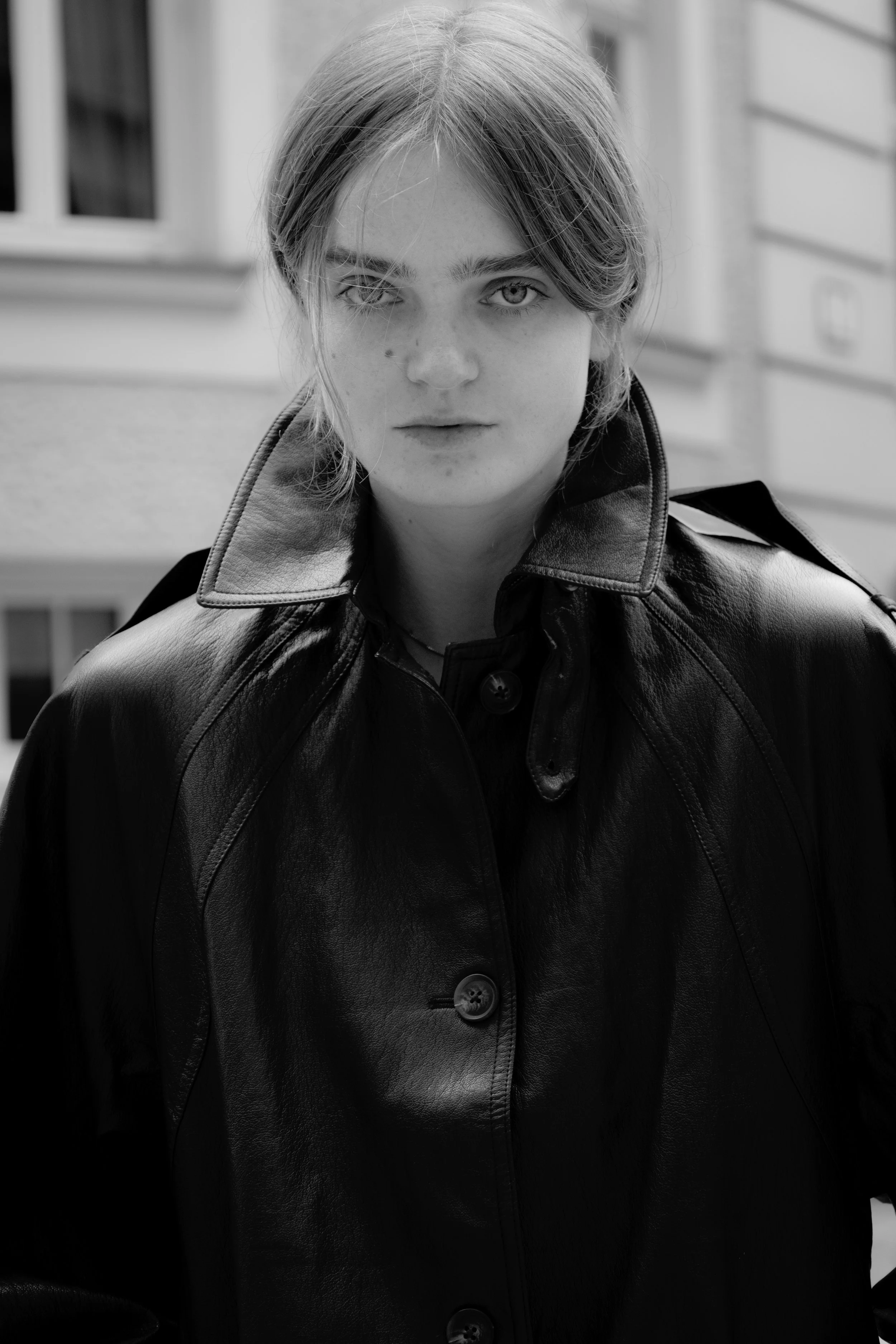 Black and white photo of a person with shoulder-length hair wearing a leather jacket, standing outdoors with buildings in the background.