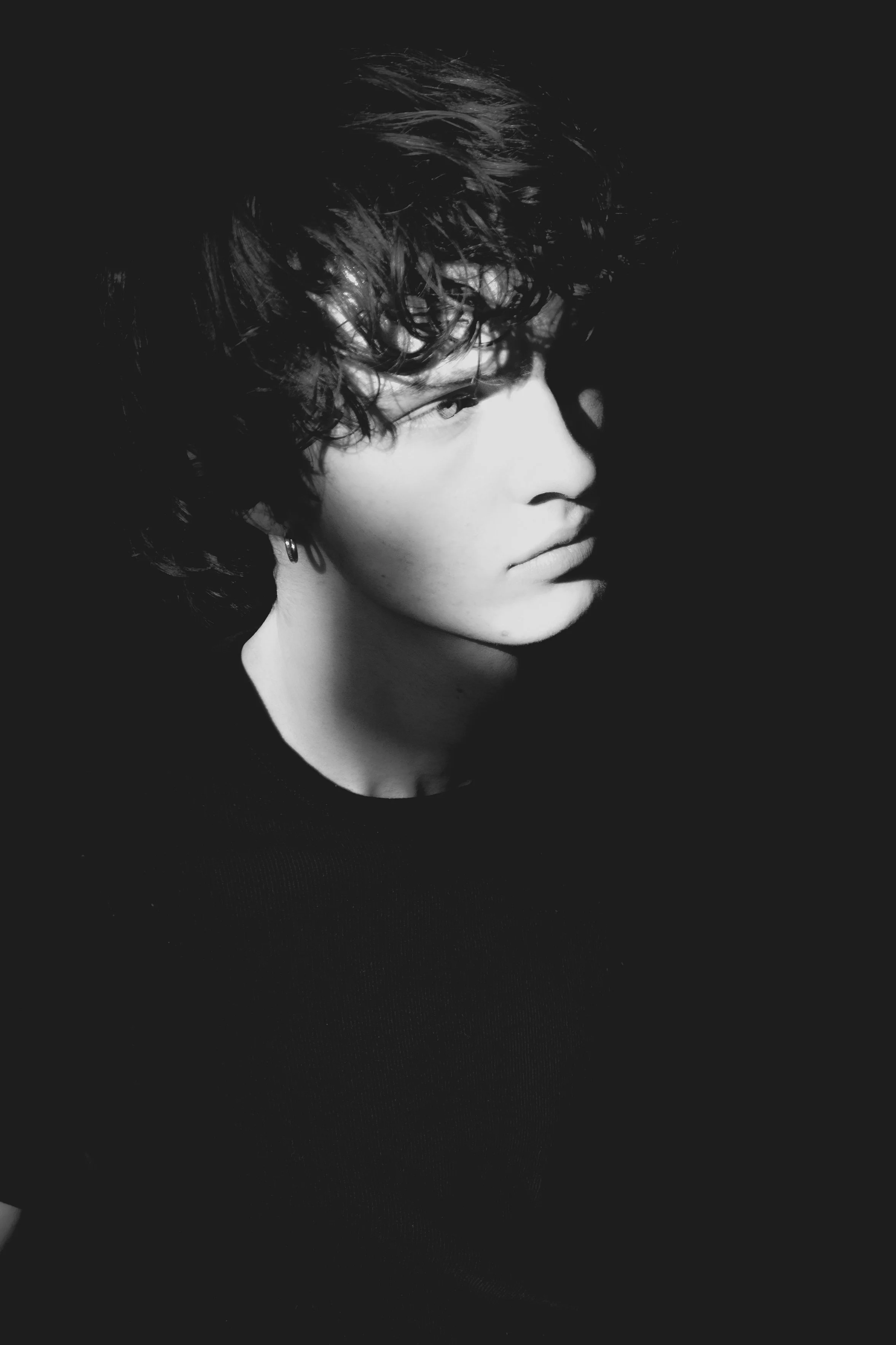 Black and white photo of a young person with curly hair, wearing a dark shirt, facing to the side with a serious expression.