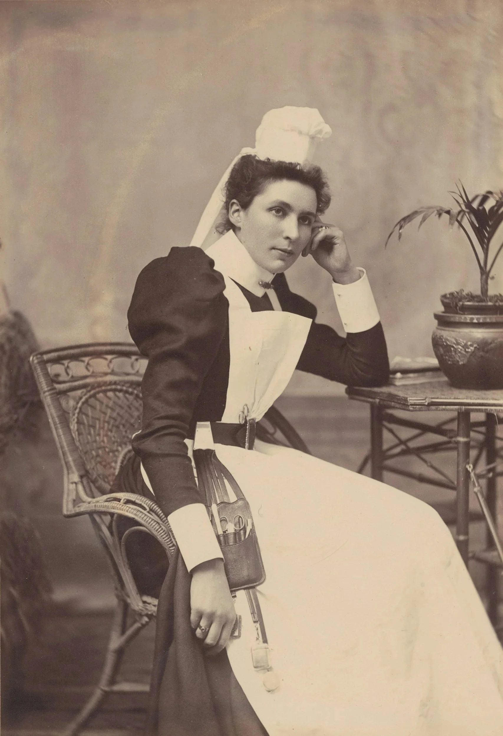 A vintage black-and-white photograph of a woman in a nurse's uniform, seated beside a table with a potted plant, holding a pair of scissors and looking at the camera.