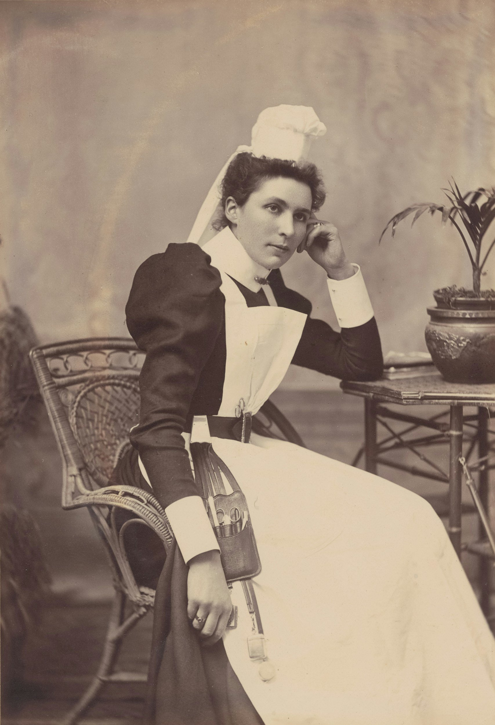 A woman dressed as a nurse sitting at a table with a potted plant, in a vintage black-and-white photograph.