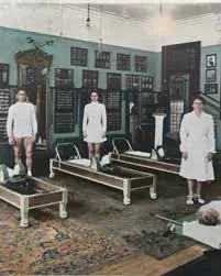 A vintage black-and-white photo of four women in white dresses standing in a room with old-fashioned furniture and equipment, possibly a hospital or clinic.