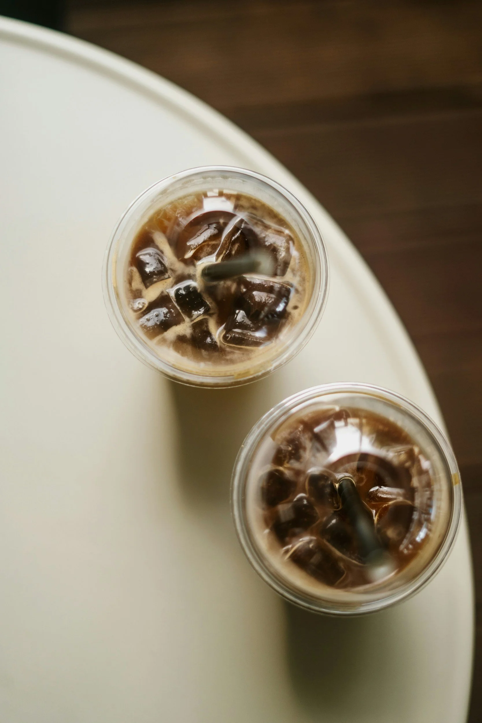 Two plastic cups filled with iced coffee and ice cubes, viewed from above on a white surface with a wooden background.