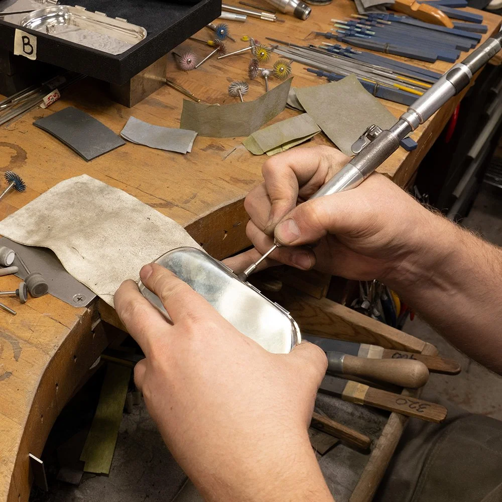 A person using a precision tool to work on a shiny silver object, likely in a jewelry or fine metalwork workshop, with various small abrasives and tools scattered on a wooden workbench.