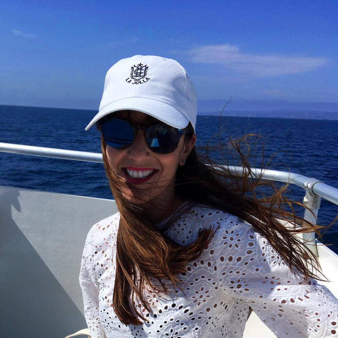 Image of Et Collectice LLC founder, Christy, smiling with a white hat and dark sunglasses on. Behind her is a railing and the blue ocean and skyline