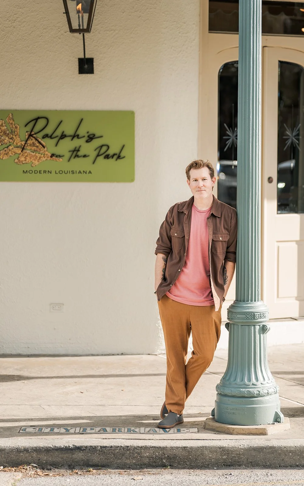 A man leaning against a green streetlamp outside a building with a sign that reads 'Ralphie in the Park, Modern Louisiana.' He is wearing a brown jacket over a pink shirt and tan pants, standing on a sidewalk.