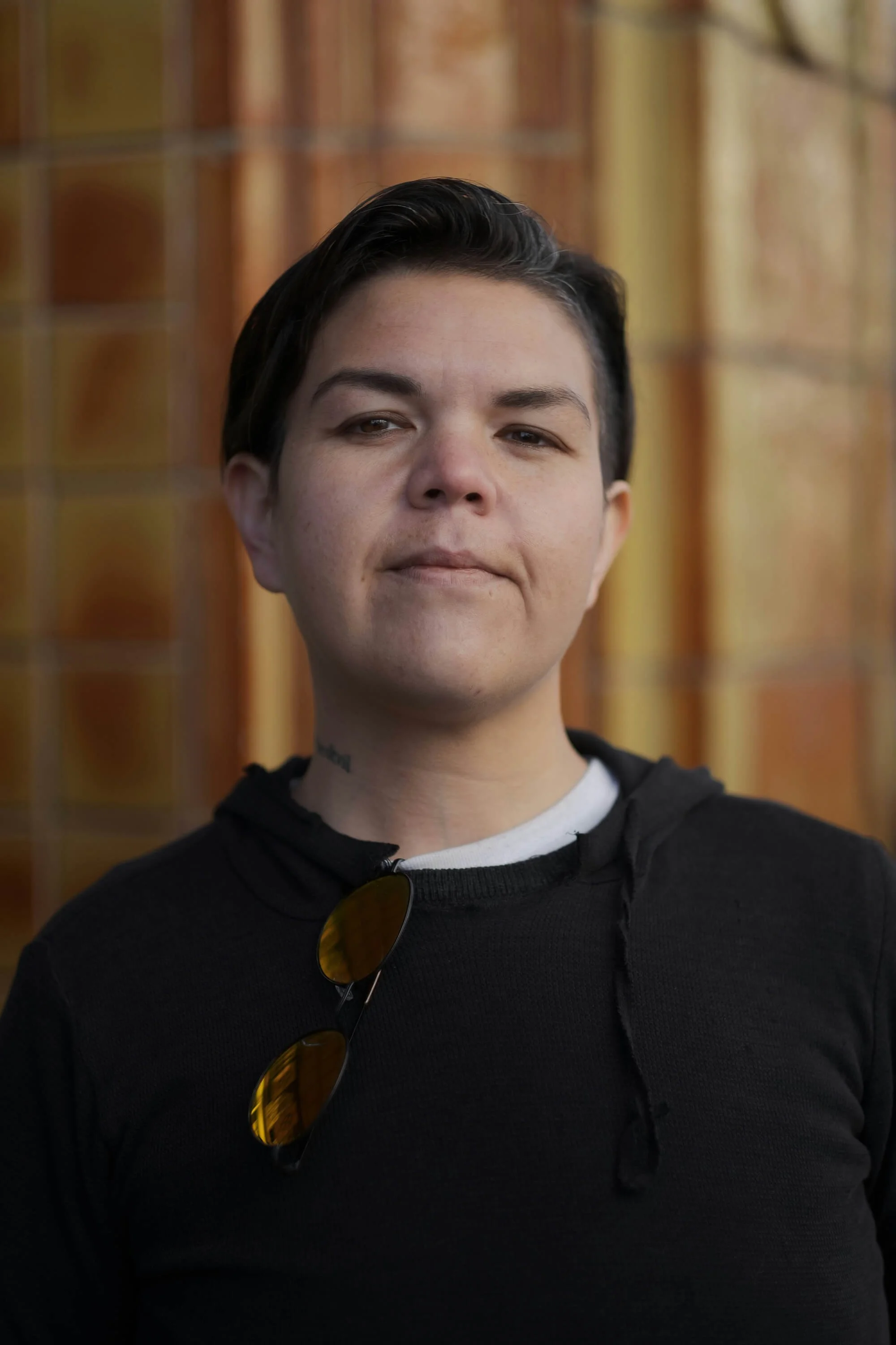 A person with short dark hair, light skin, and a slight smile, wearing a black hoodie and sunglasses hanging from the hoodie, standing in front of a blurred wooden background.