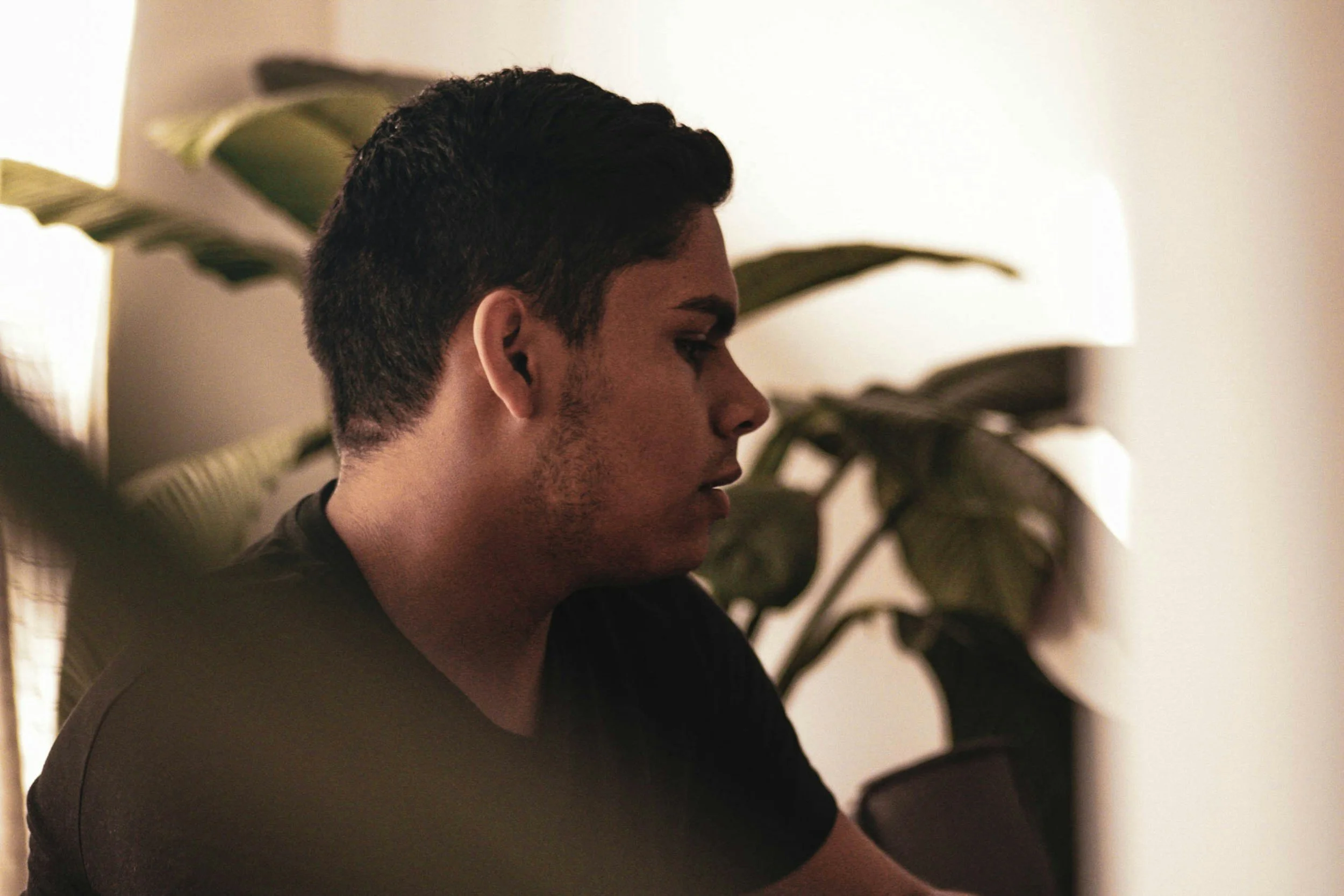 Side profile of a young man with short dark hair, wearing a black shirt, sitting indoors near a large green leafy plant.