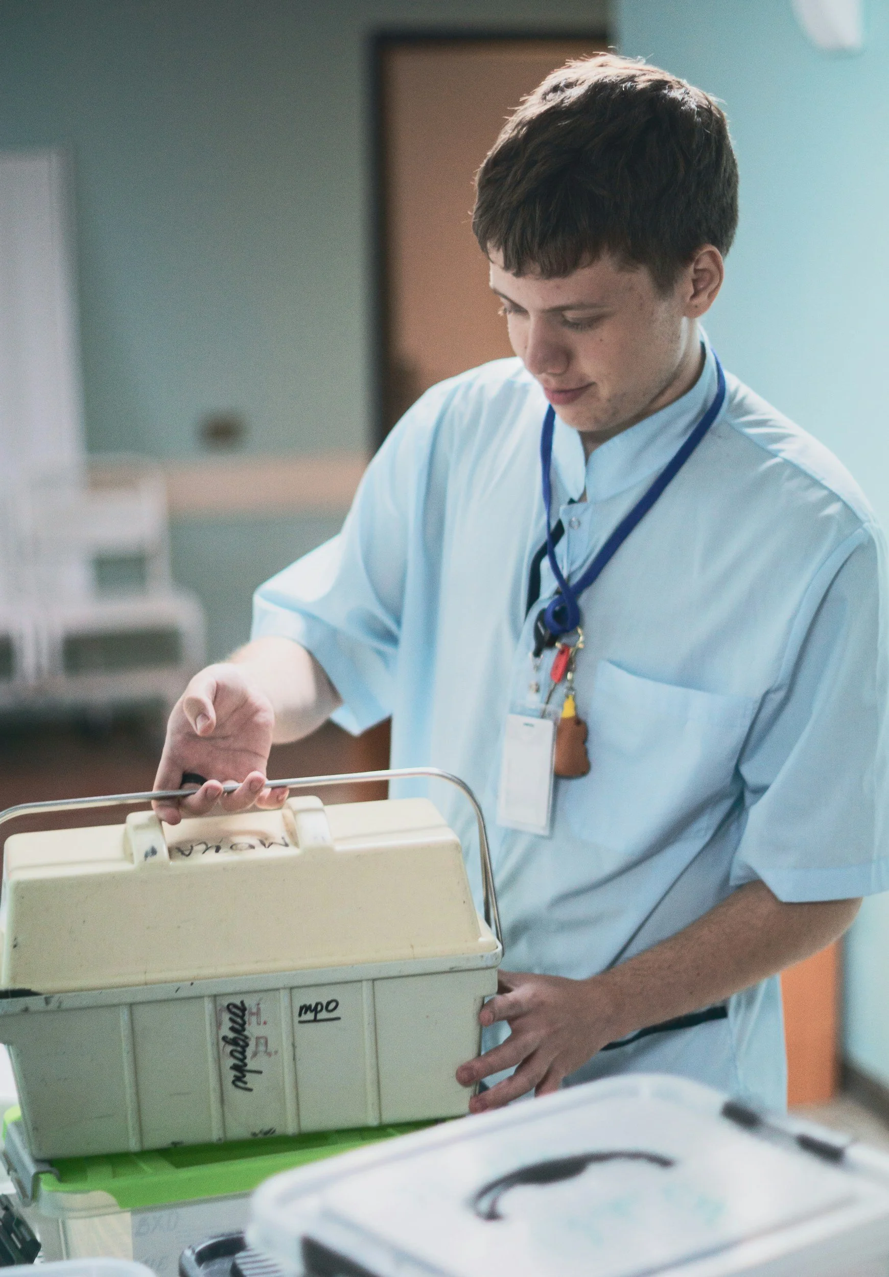 A young male healthcare professional with short brown hair and light skin, wearing light blue scrubs and a stethoscope around his neck, is working with a medical machine in a hospital or clinic setting.