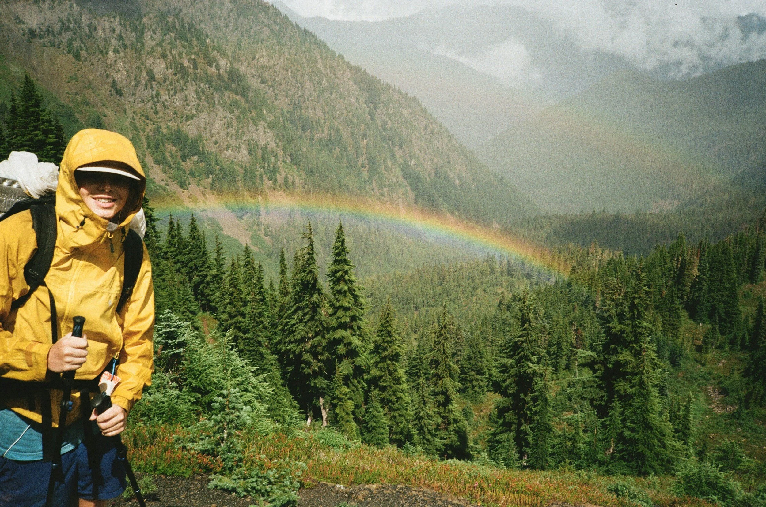 Person in yellow rain jacket and cap with backpack standing in lush green forest with rainbow in the background mountain landscape