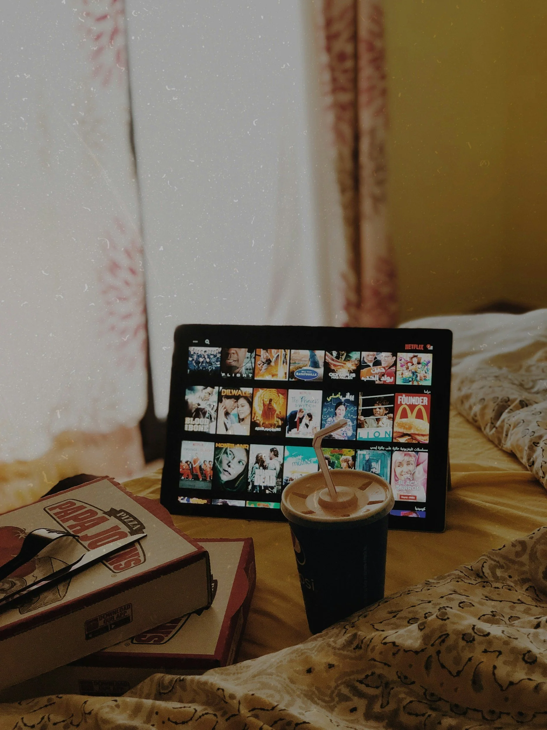 A tablet displaying a Netflix interface with various movies and shows, a black cup with a straw in front of it, a Pizza Hut box, and a Domino's box on a bed with a yellow blanket and leopard print sheets.